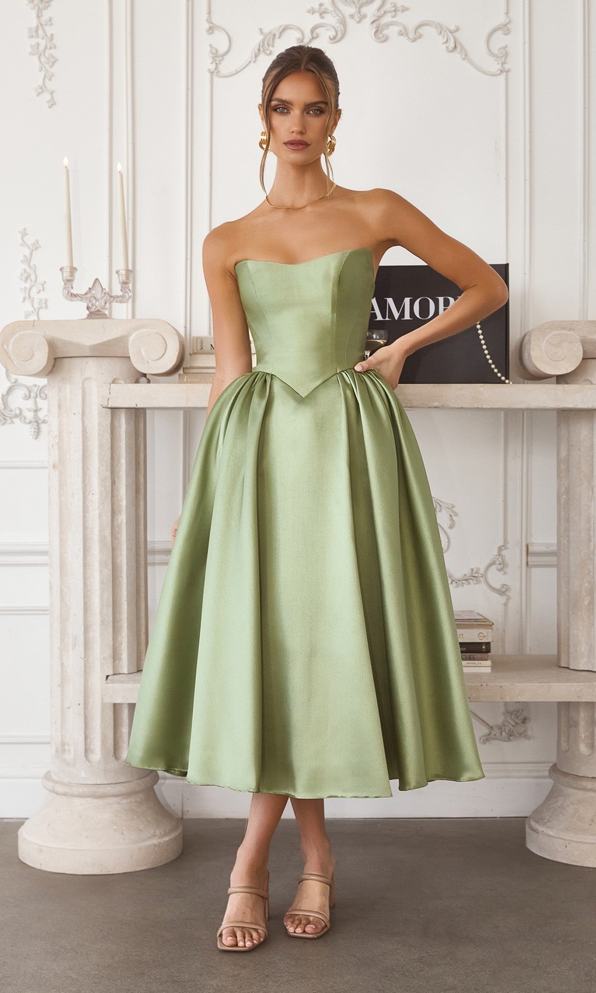 A woman stands indoors wearing the Amoris T822 midi prom dress in sage, featuring a strapless fitted bodice and elegant A-line skirt, paired with beige sandals.