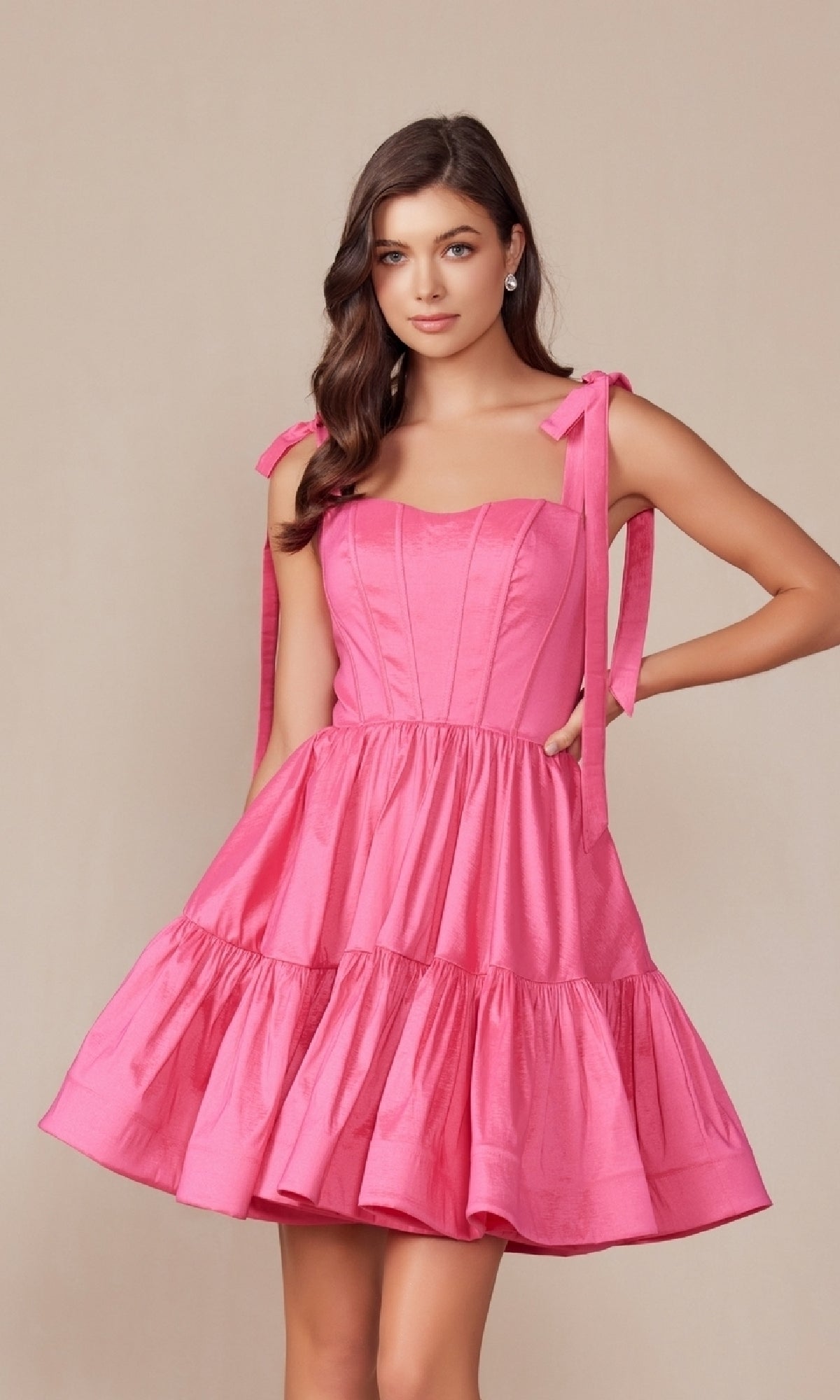 A woman wears the Shoulder-Tie Short Lace-Up A-Line Hoco Dress T869, a sleeveless pink party dress with tied shoulders and flared skirt, standing against a plain beige background.