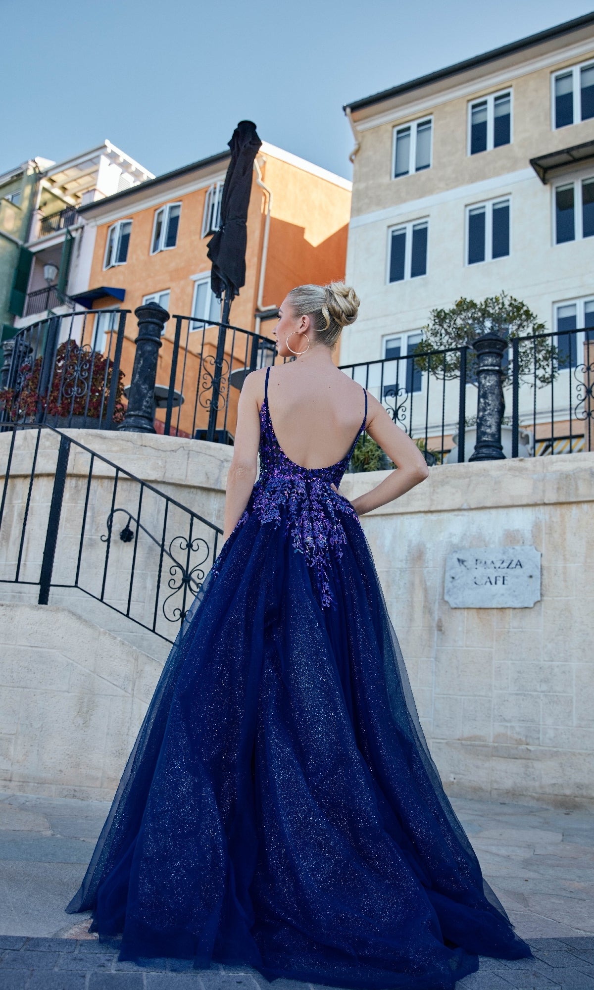 Wearing the Tina Holly TE216B long formal dress in dark blue, a woman stands outdoors on a stone path, facing away, with buildings and stairs in the background. The gown features a floor-length design and high-leg slit.