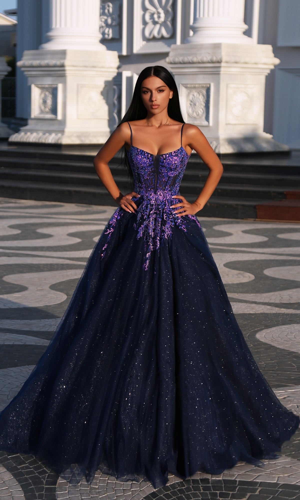 A woman stands outside a white classical building wearing the Tina Holly TE216B, a dark blue glitter tulle ball gown with purple floral appliques and thin straps.