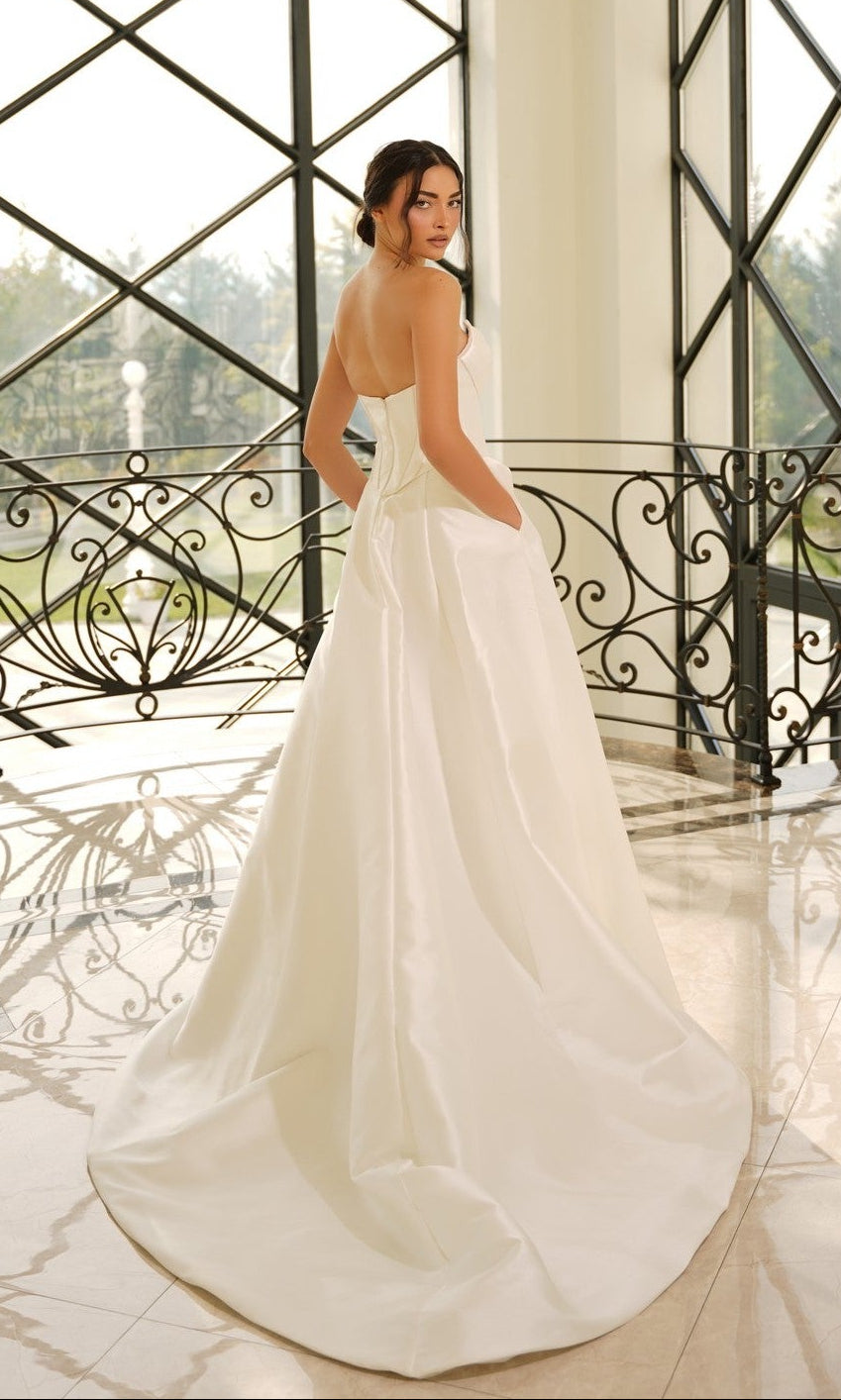 A woman wearing the Strapless Mikado Wedding Dress: Tina Holly TE704 with pockets stands indoors before large windows and ornate railings, glancing over her shoulder.