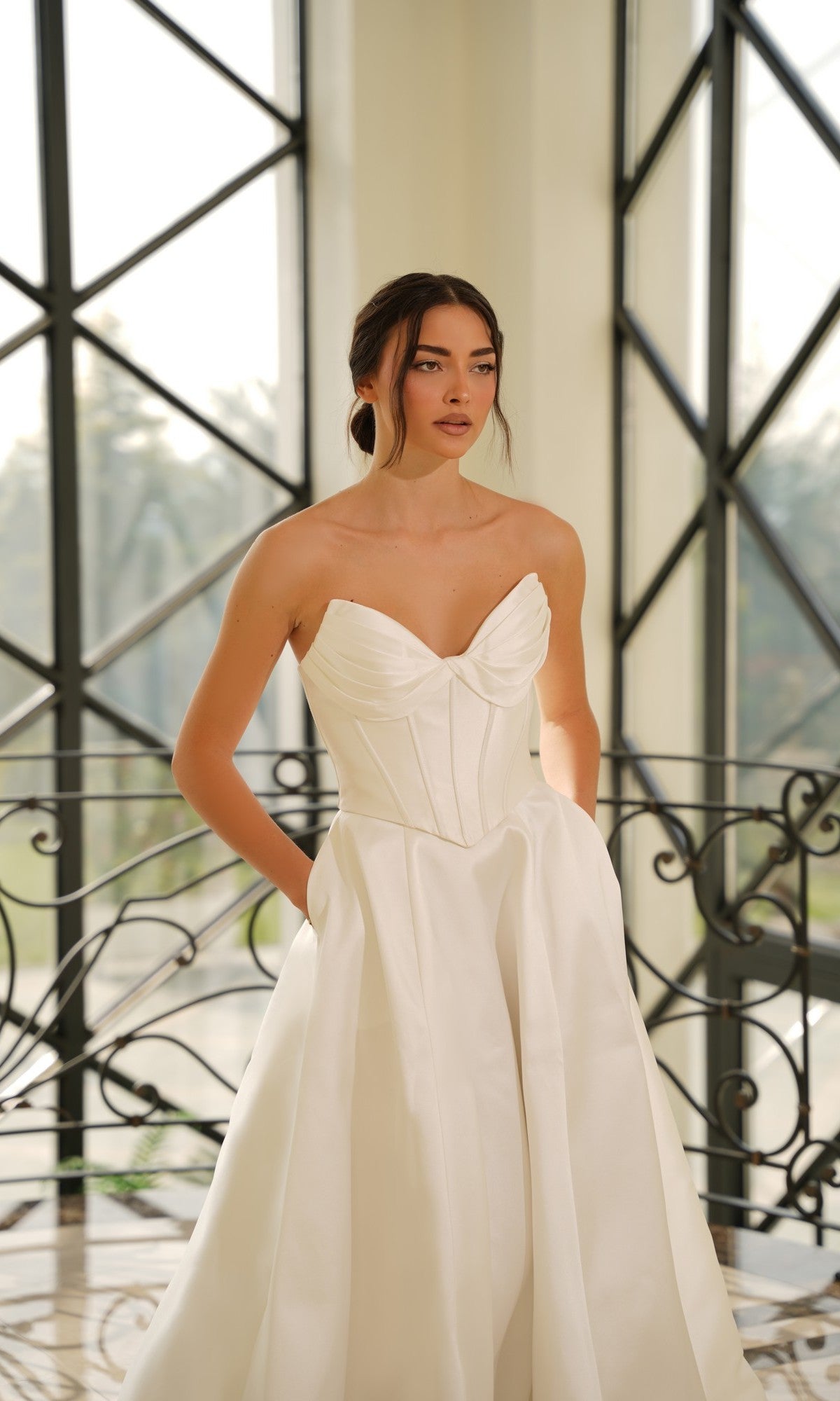 A woman in the Strapless Mikado Wedding Dress: Tina Holly TE704 stands indoors by large windows and decorative iron railings, gazing slightly to the side.
