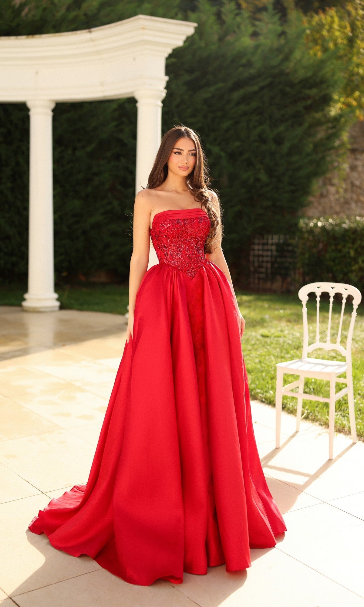 A woman in the Tina Holly TE706 long formal dress, featuring a strapless embroidered corset bodice, stands on a patio by a white chair, with greenery and white columns in the background.