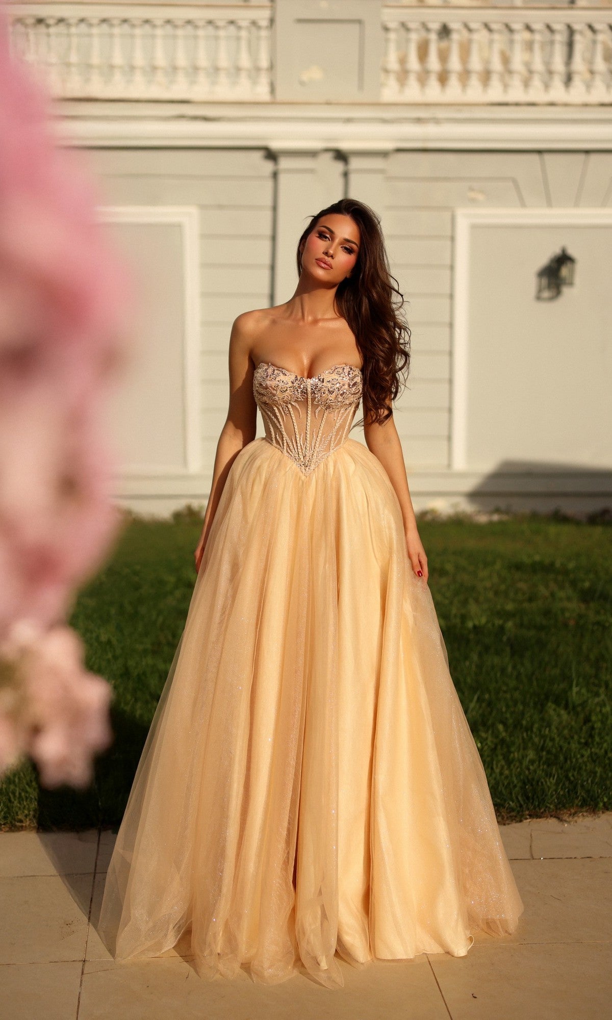 A woman in the champagne Tina Holly TE708 long formal dress, featuring a strapless glitter ball gown silhouette with a fitted sheer corset bodice, stands outdoors in front of a light gray building.