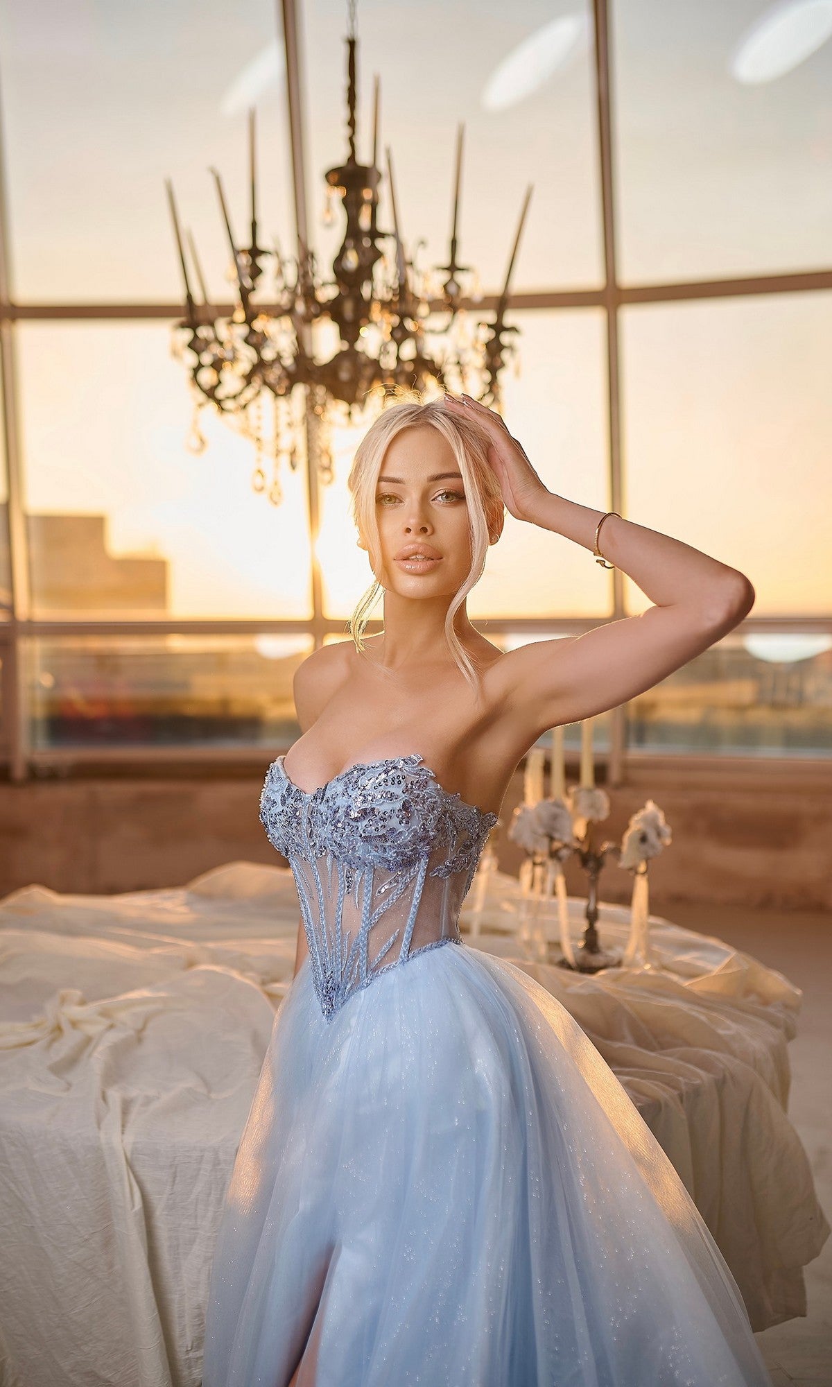 A woman in the Tina Holly TE708 long formal dress—a pale blue strapless glitter ball gown with a lace corset bodice—stands indoors by a table, under a sparkling chandelier and sunset-lit windows.