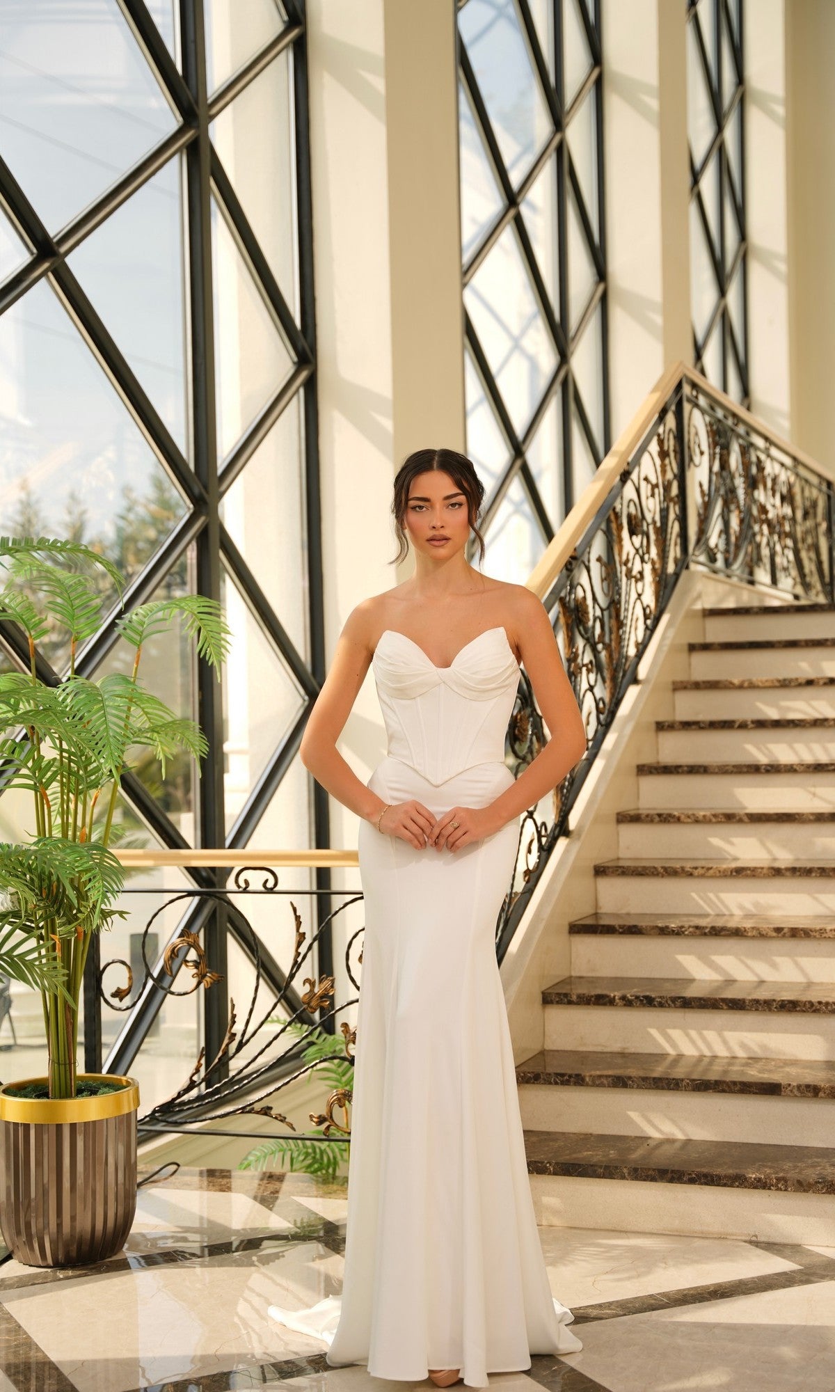 A woman wearing the Contemporary Wedding Dress: Tina Holly TE709 stands at the base of a staircase with large windows and a decorative railing in the background.