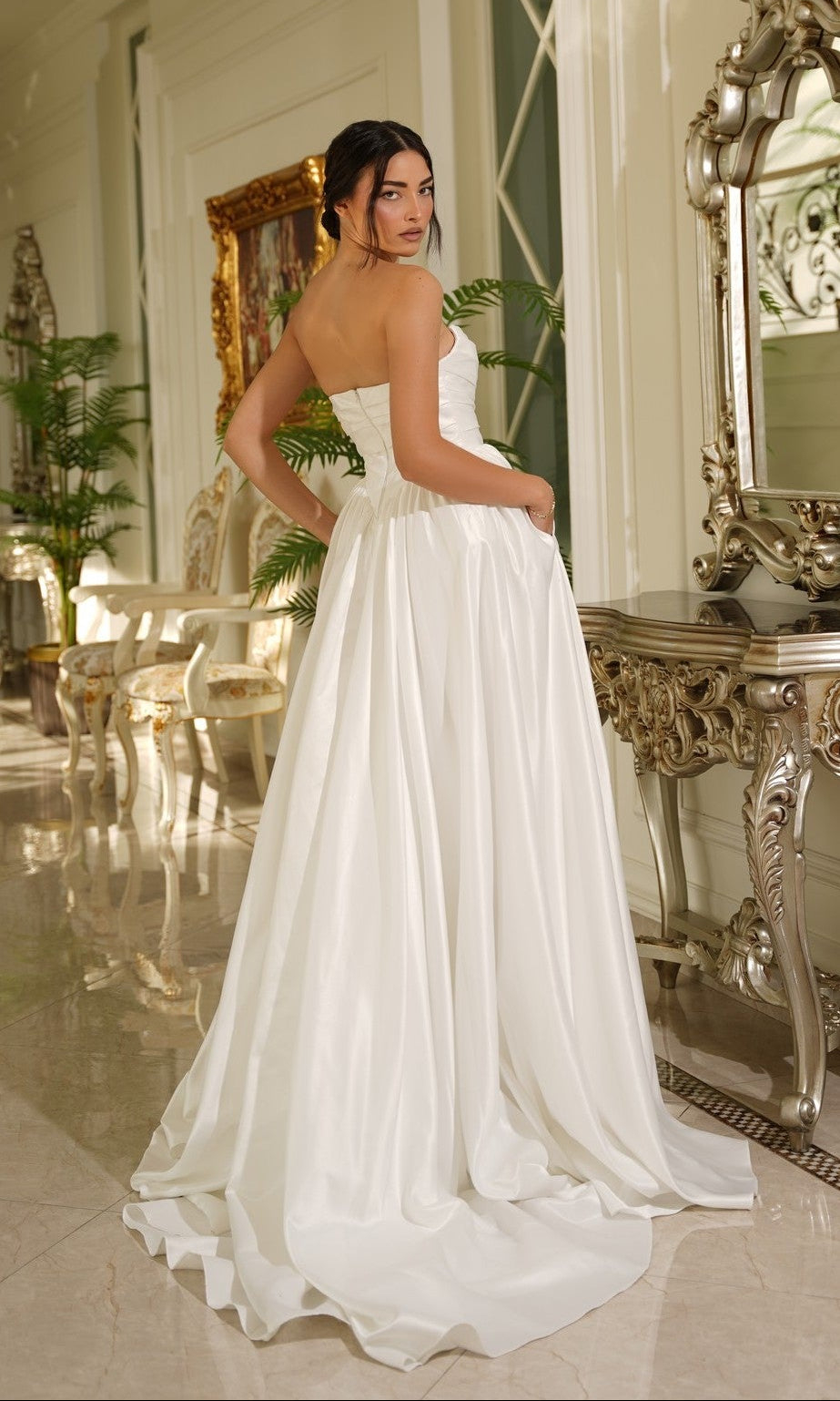 A woman in the Long Off-White Satin Dress: Tina Holly TE810 stands indoors near ornate furniture, facing away while glancing over her shoulder.