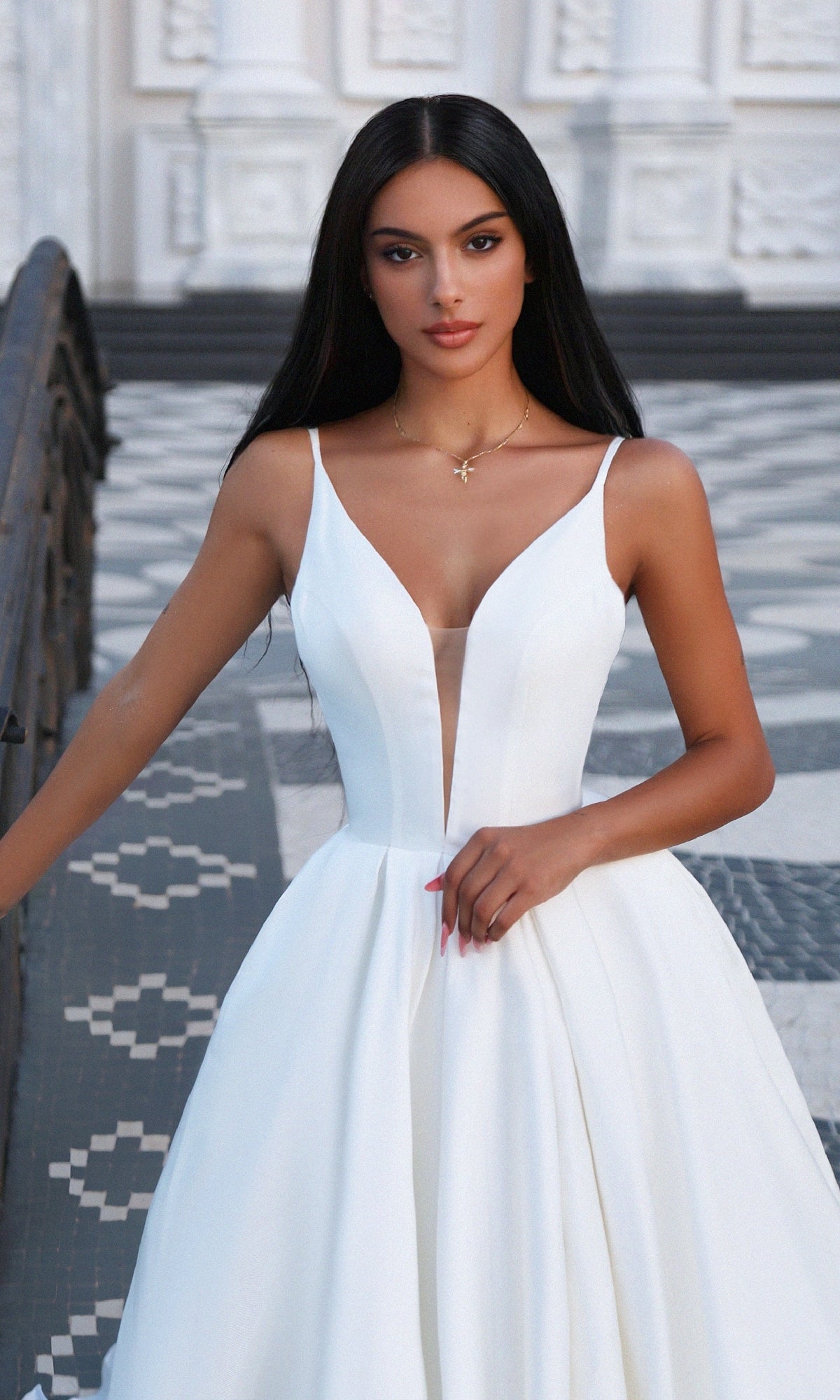 A woman wears the Off-White Mikado Prom Ball Gown: Tina Holly TF002, featuring a long silhouette and deep V-neckline, as she stands on patterned tiles by a railing against a white architectural backdrop.