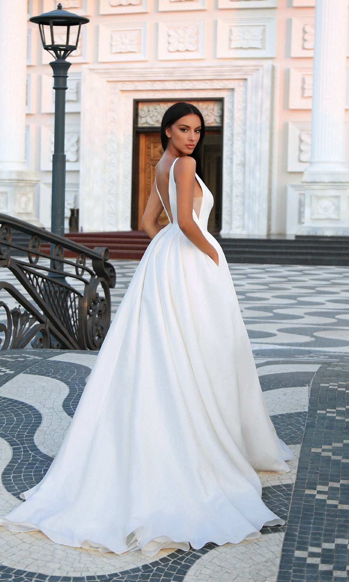 A woman wearing the Off-White Mikado Prom Ball Gown: Tina Holly TF002 stands outdoors on a patterned stone walkway near a staircase and an ornate building façade.
