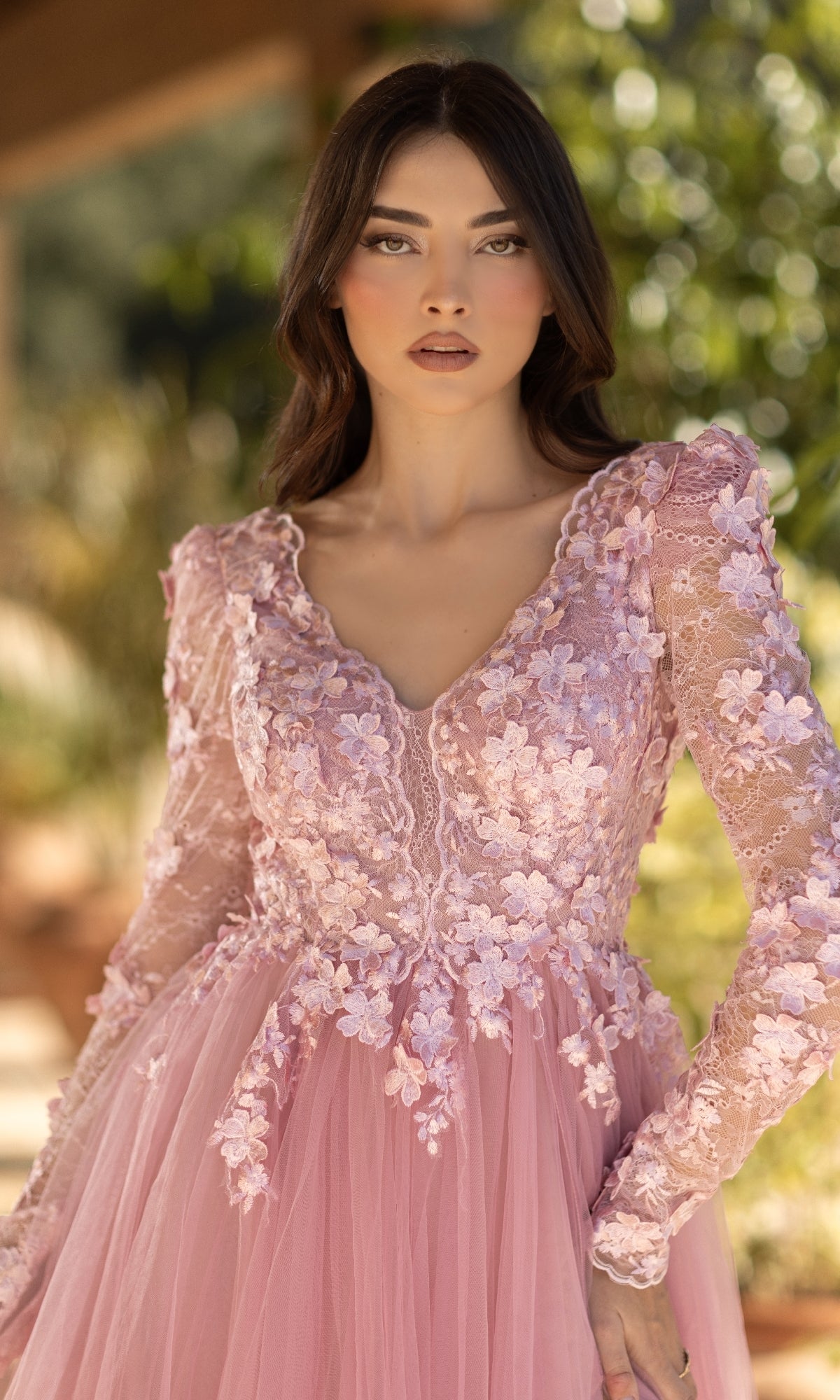 A woman stands outdoors amid greenery, wearing the Long Pink Formal Dress: Tina Holly TF318F featuring a pastel pink hue, lace bodice, and floral embroidery.