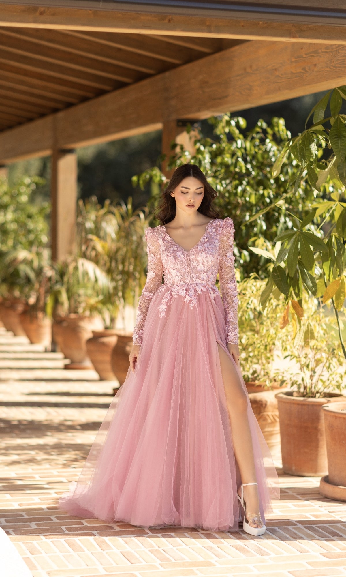 A woman in the Long Pink Formal Dress: Tina Holly TF318F, featuring a lace bodice and high slit, stands on a sunlit outdoor walkway lined with potted plants.