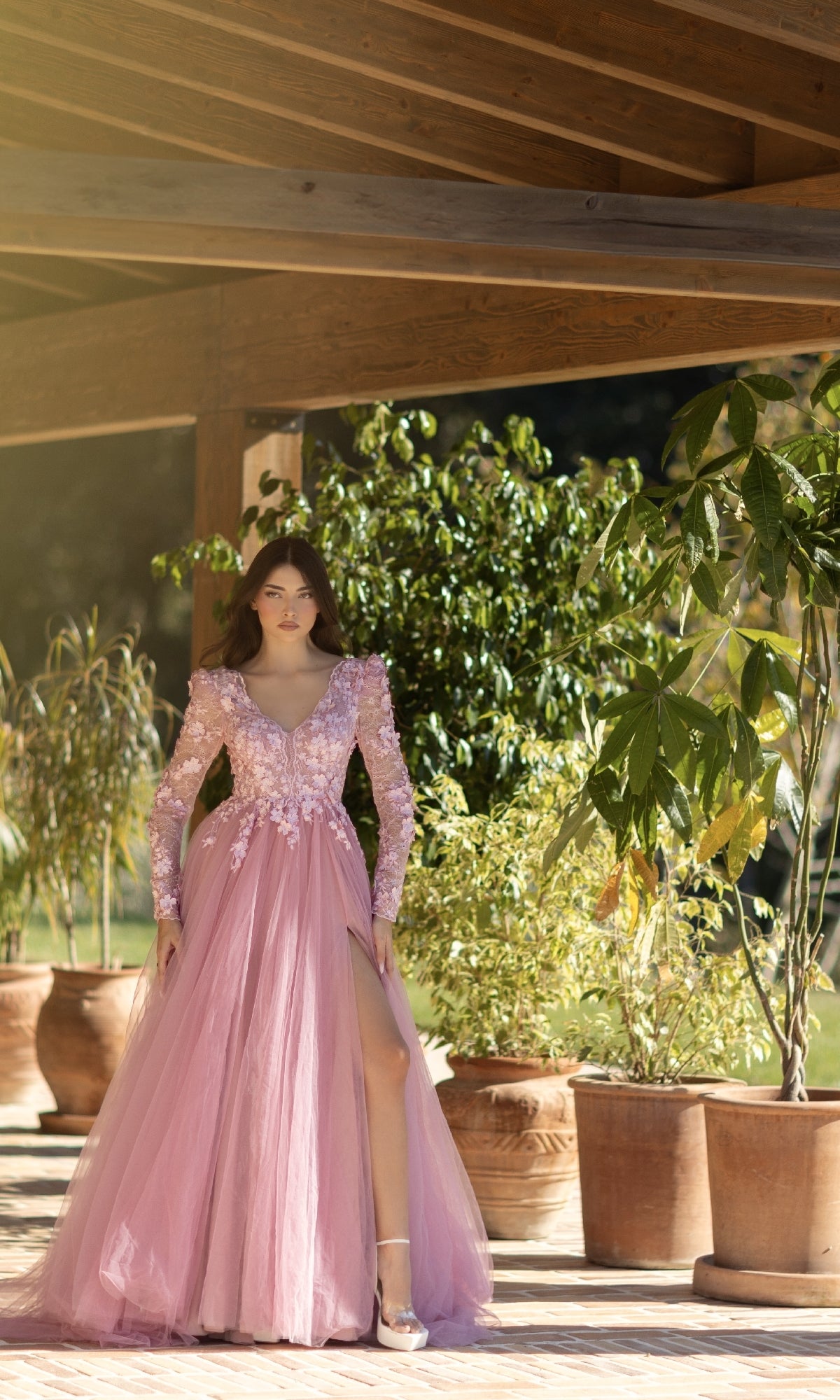 A woman in the Long Pink Formal Dress: Tina Holly TF318F, featuring a lace bodice and high slit, stands under a wooden pergola surrounded by potted plants.