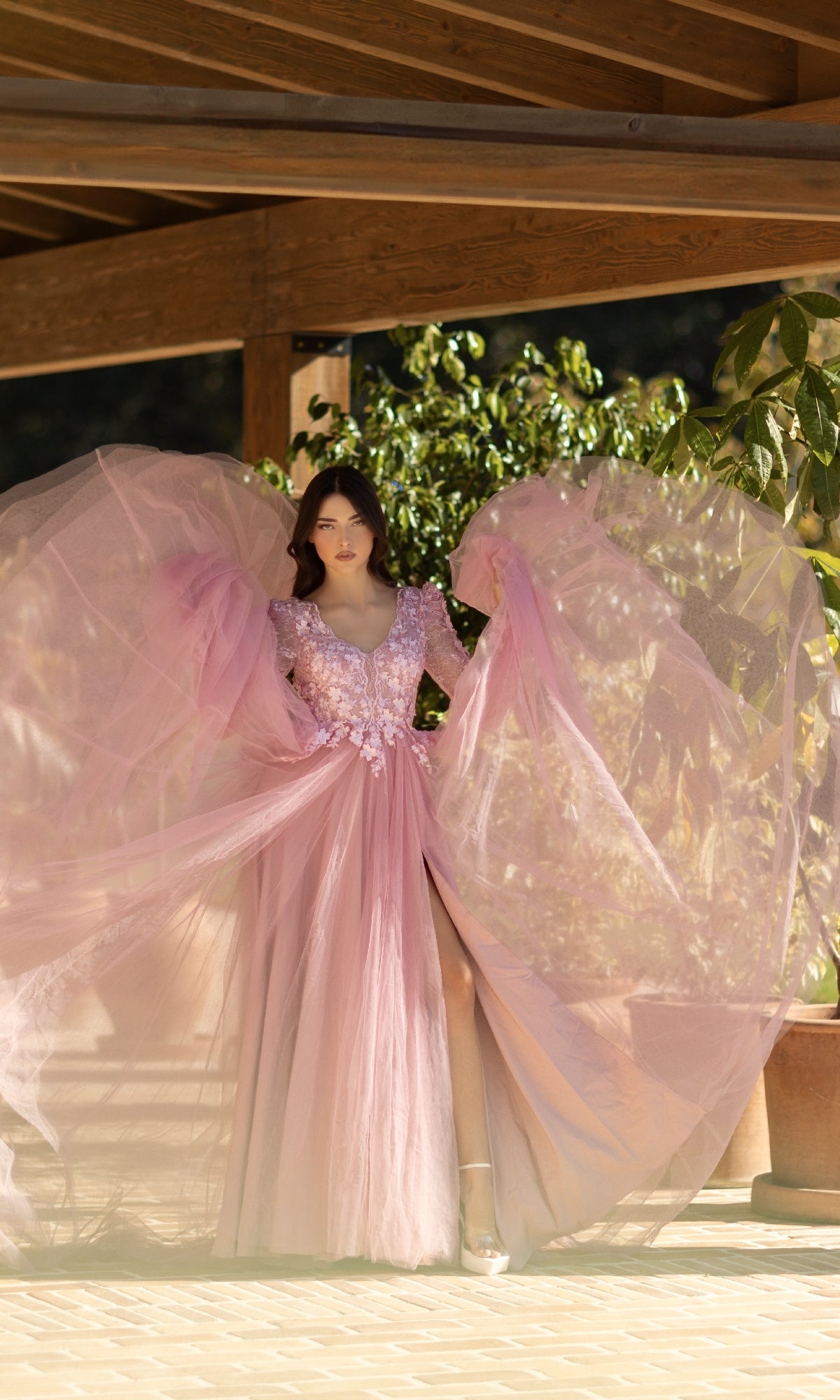 A woman poses gracefully under a wooden pergola in the Long Pink Formal Dress: Tina Holly TF318F, featuring sheer wide sleeves, floral embellishments, and an elegant lace bodice, surrounded by potted plants.