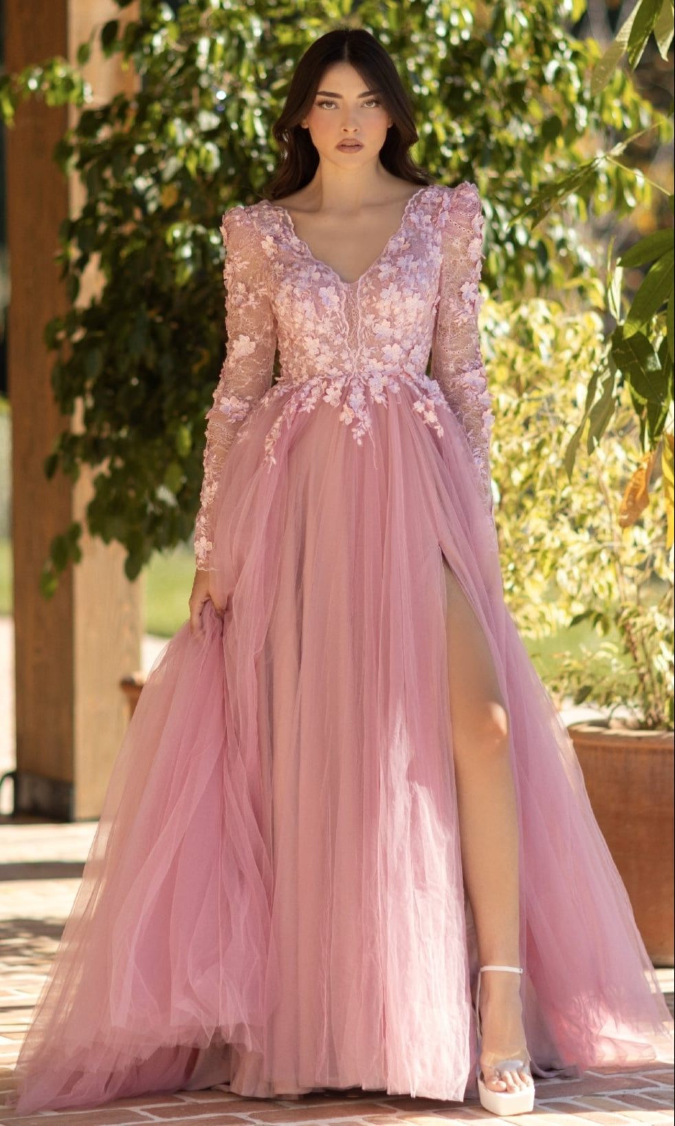 A woman stands outdoors wearing the Long Pink Formal Dress: Tina Holly TF318F, featuring floral embroidery, a lace bodice, sheer long sleeves, a plunging neckline, and a thigh-high slit.