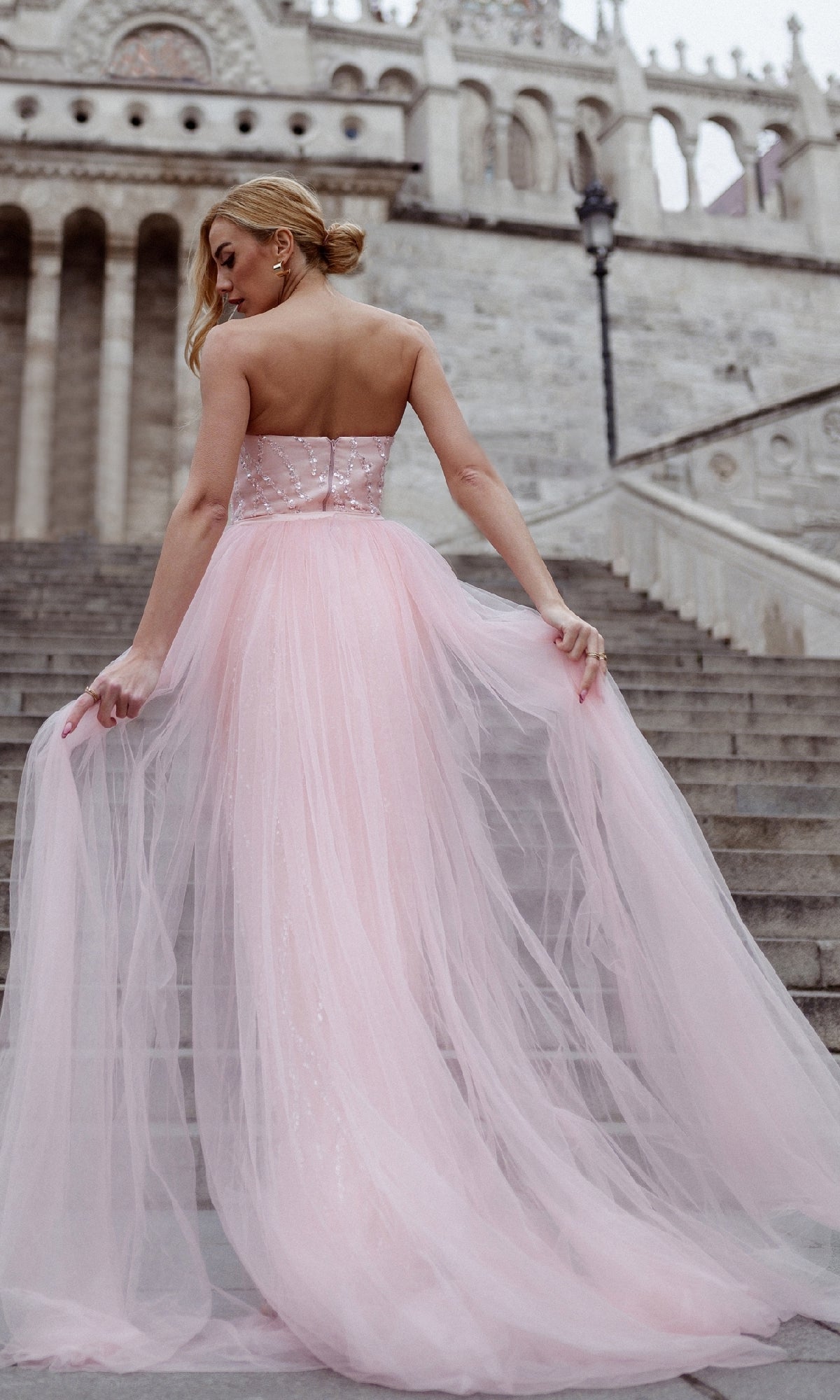 A woman in the Overskirt Strapless Prom Gown: Tina Holly TK118, a light pink tulle evening dress, stands on stone steps outside a grand building, facing away from the camera.