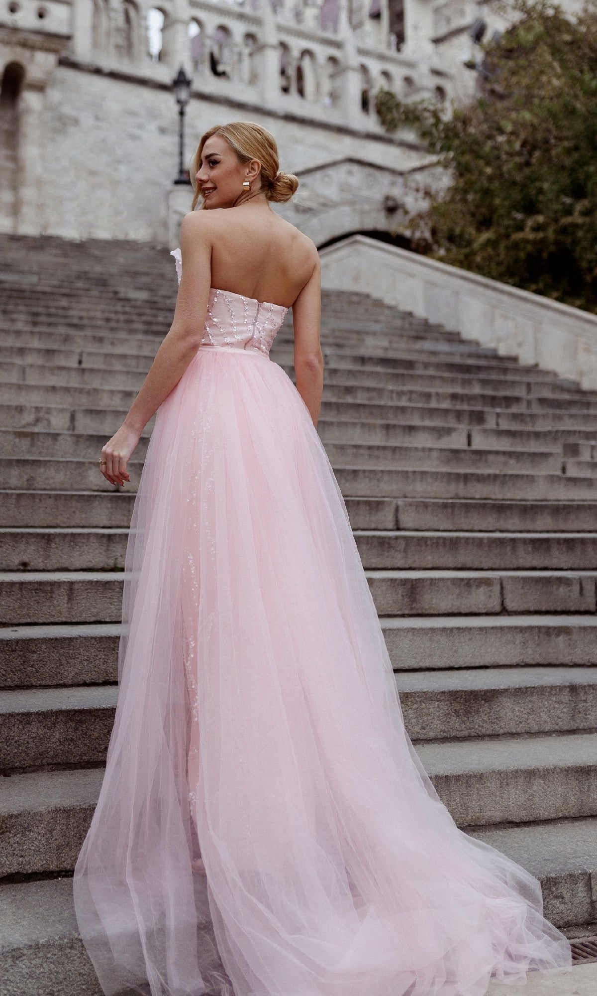 A woman in the Overskirt Strapless Prom Gown: Tina Holly TK118 stands on stone steps, looking over her shoulder.