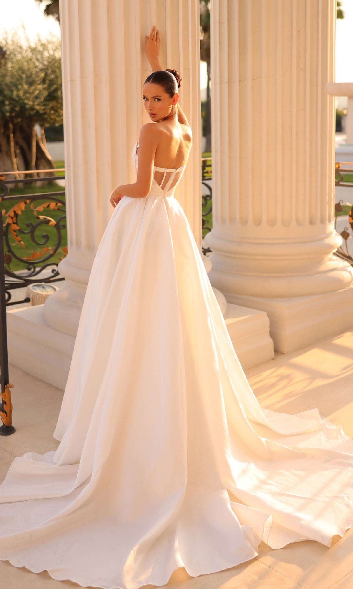 A woman in the Strapless Off-White Formal Dress: Tina Holly TK241, featuring a long train, stands before large white columns and looks over her shoulder.