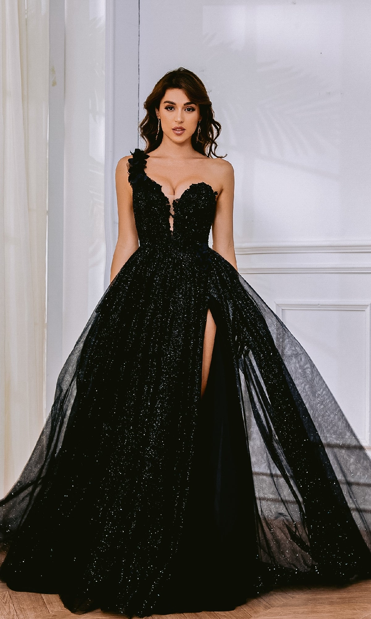 A woman stands indoors wearing the One-Shoulder Glitter Prom Gown: Tina Holly TK554, a floor-length black dress with a fitted bodice, thigh-high slit, and sparkling one-shoulder design.