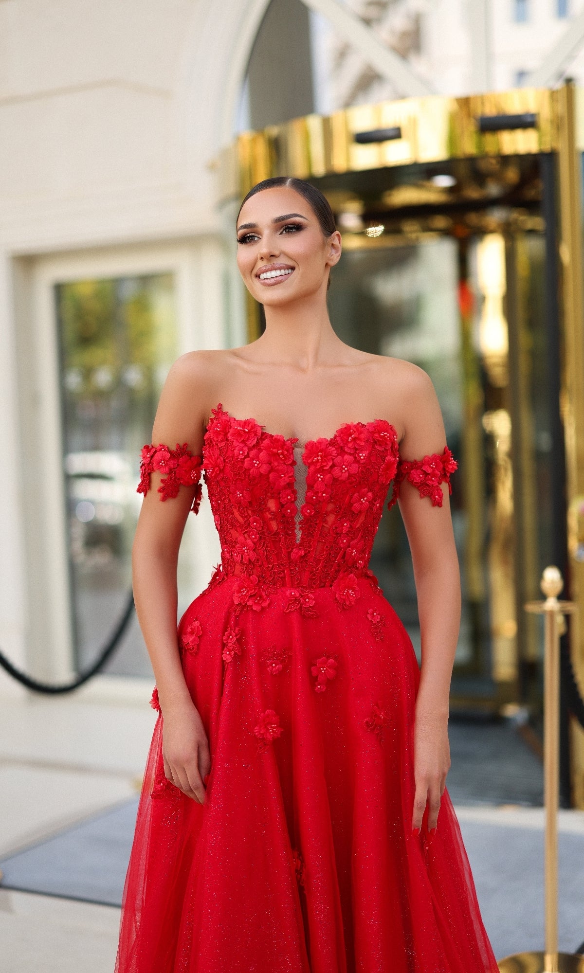 A woman stands outside a building, smiling at the camera in the Tina Holly TK582 long prom dress—an off-the-shoulder, floor-length red gown with a corset bodice and 3D flower appliques.
