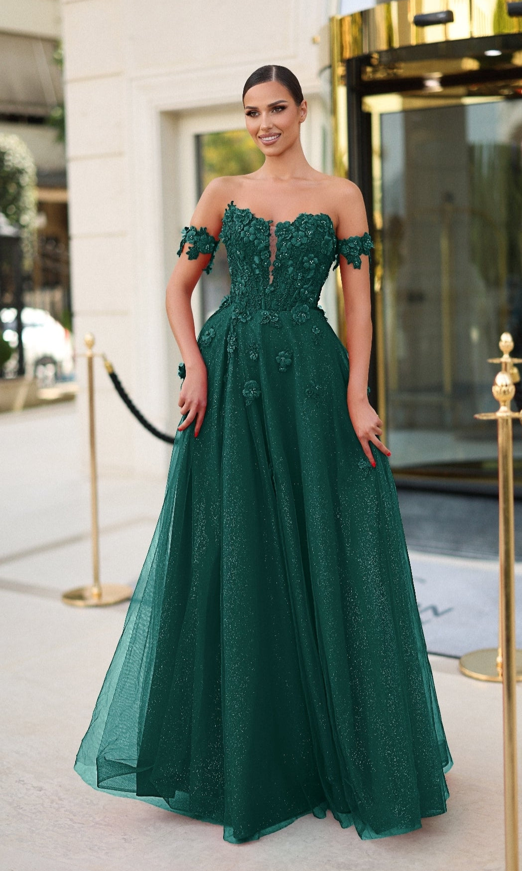 A woman stands outside a building wearing the Tina Holly TK582, an off-the-shoulder, floor-length green prom dress featuring 3D flower appliques and a sheer bodice.