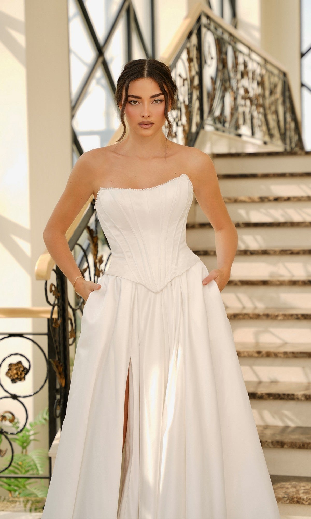 A woman wears the Tina Holly TK803 Off-White Satin Wedding Dress, featuring a strapless corset bodice and pockets, as she stands indoors before a decorative staircase.