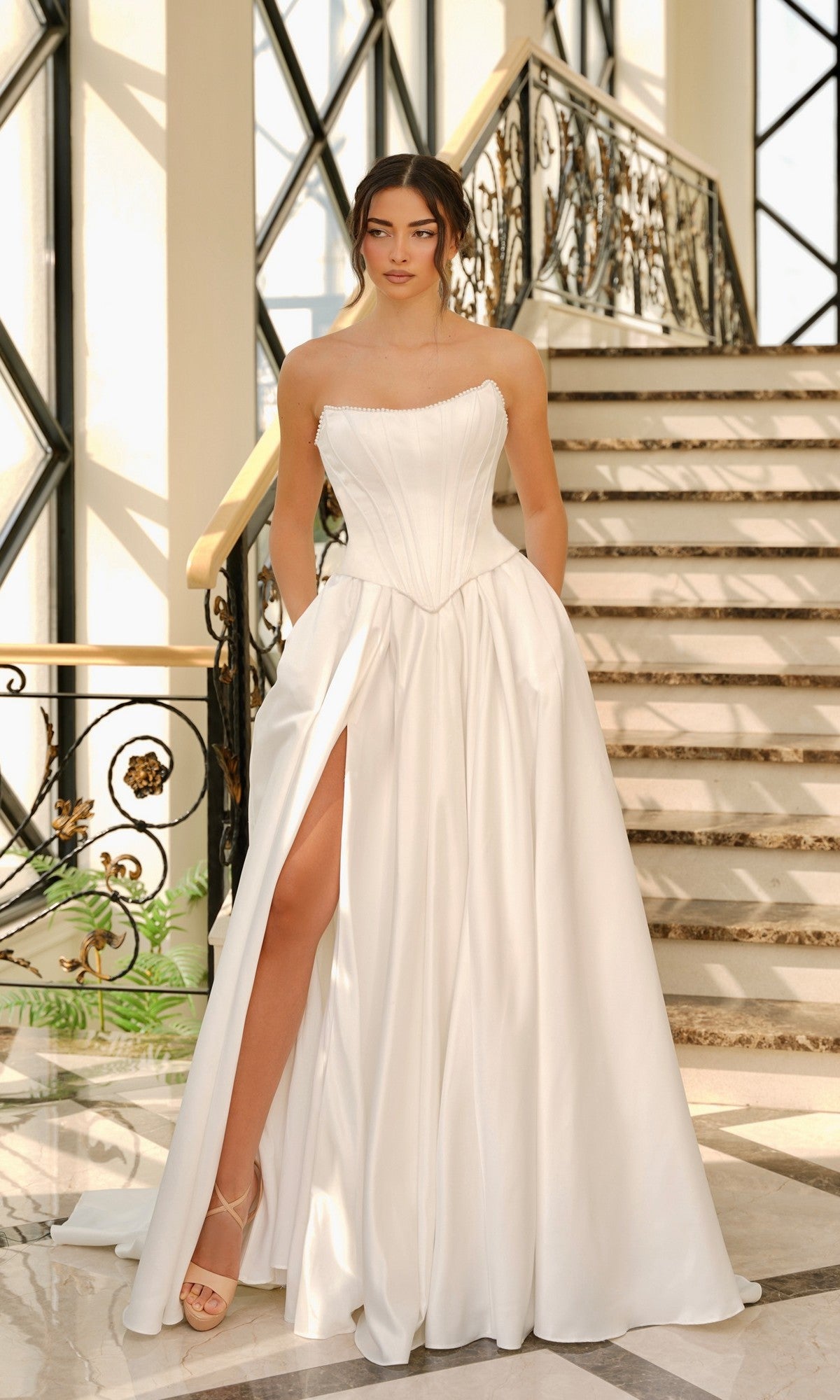 A woman poses indoors in front of a decorative staircase, wearing the Off-White Satin Wedding Dress: Tina Holly TK803, featuring a strapless design, structured corset bodice, and high slit.