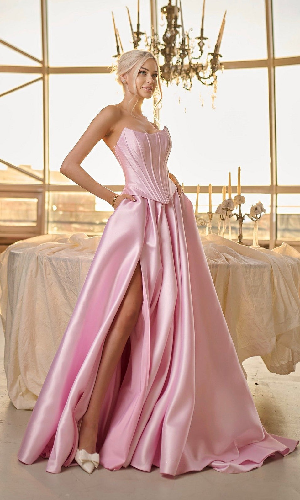 A woman in the Tina Holly TK804 long formal dress—strapless, light pink satin with a corset bodice and high slit—stands indoors by a candlelit table beneath a chandelier.