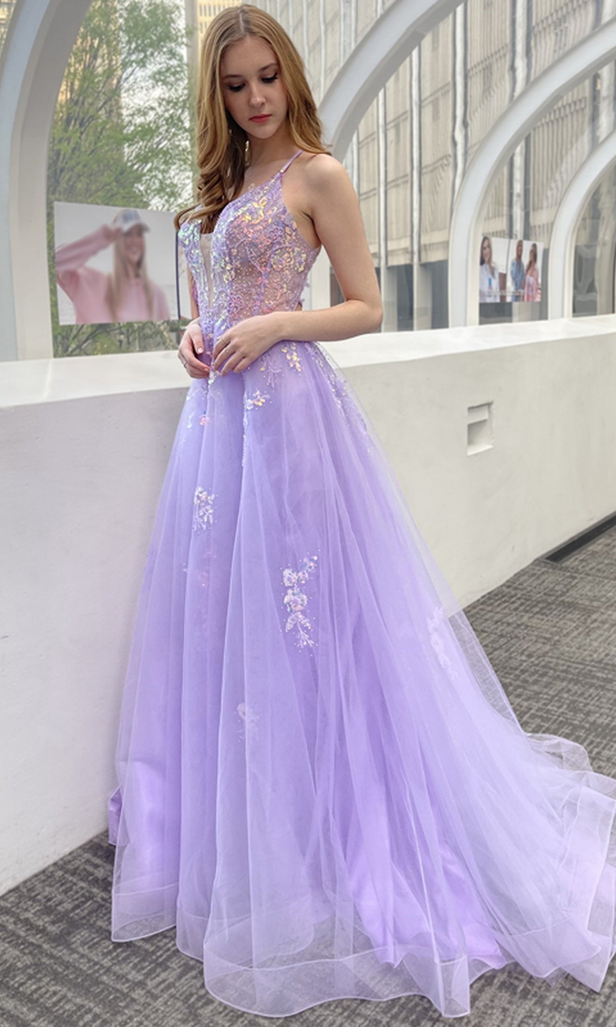 A woman wearing the Floral-Embellished Pastel Prom Ball Gown TM1003 stands indoors by a curved window, with city buildings visible outside.