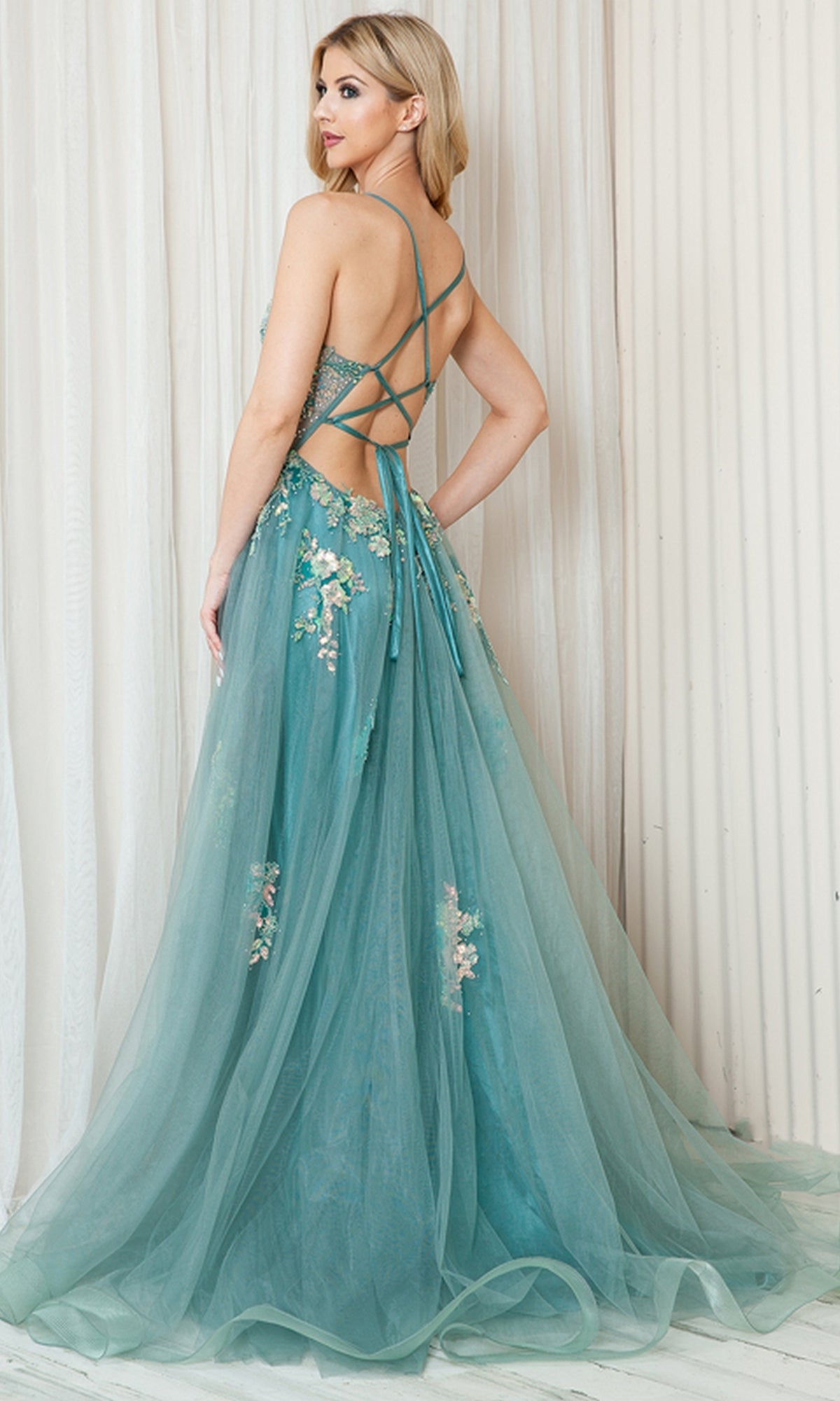 A woman in the Floral-Embellished Pastel Prom Ball Gown TM1003, featuring floor-length teal tulle, floral accents, and a crisscross open back, stands before white paneled curtains.