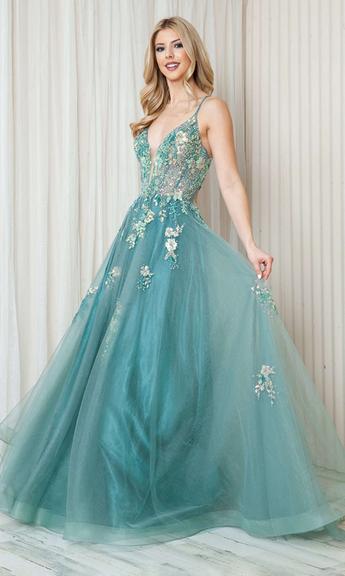 A woman poses indoors against a light, pleated backdrop wearing the Floral-Embellished Pastel Prom Ball Gown TM1003, featuring a deep V neckline and floral embellishments.