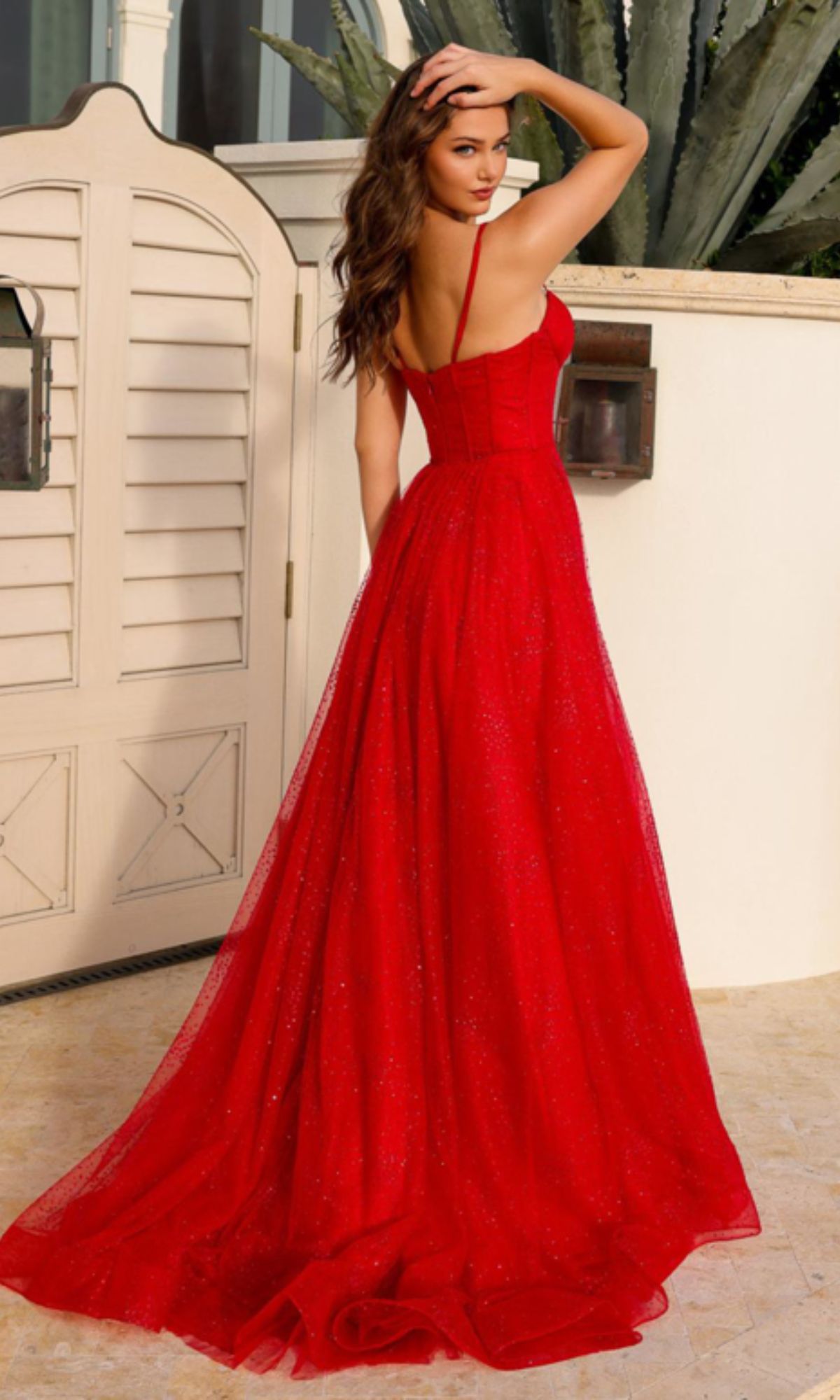 A woman stands outdoors in a sleeveless, floor-length Amelia Couture Corset Glitter Prom Dress TM1022 with thin straps, facing away and looking over her shoulder.