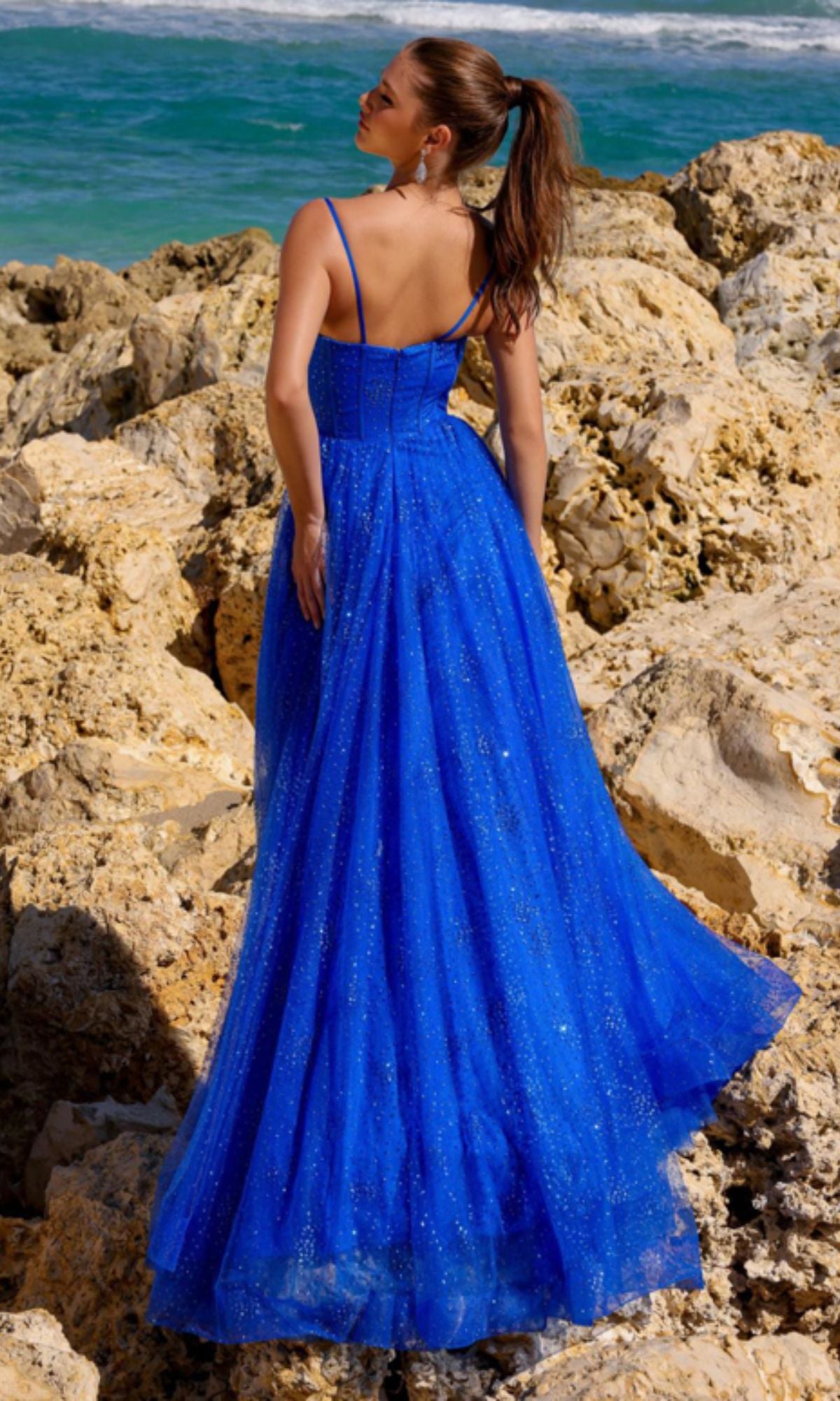 A woman in the Amelia Couture Corset Glitter Prom Dress TM1022 stands on rocky terrain by the ocean, facing away from the camera.