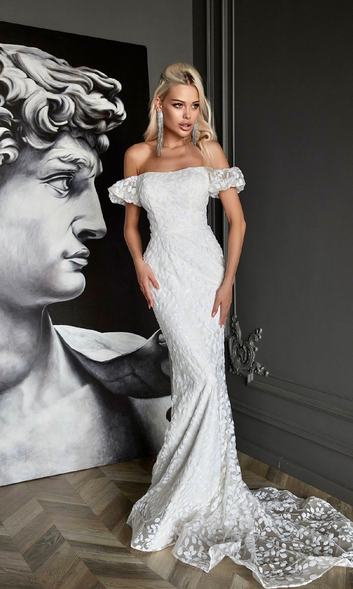 A woman wearing the Off-Shoulder White Formal Dress: Tina Holly Tk024W stands indoors beside a large grayscale painting of a classical male statue head.