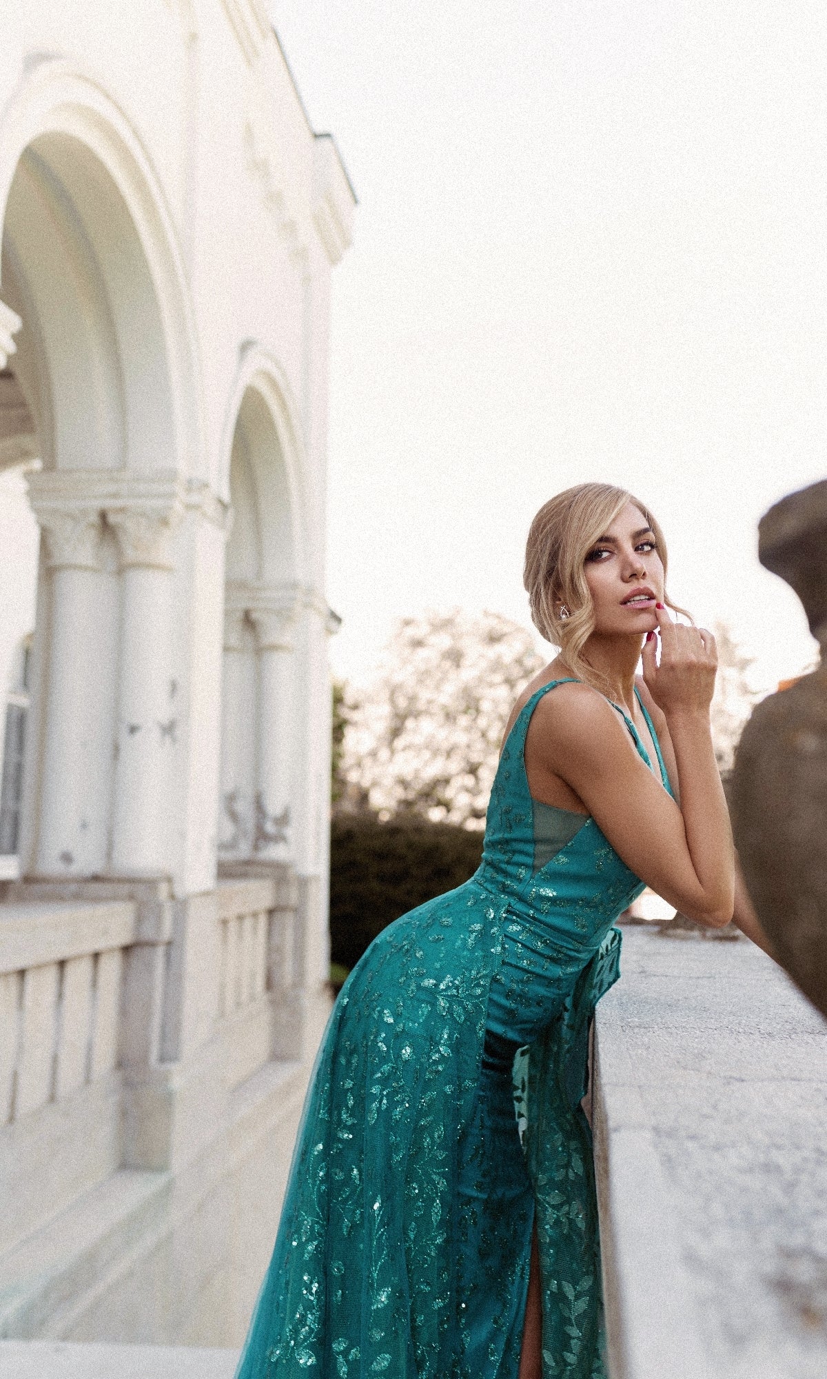 A woman in the Ruffle Overskirt Long Prom Gown: Tina Holly TW025, a teal sleeveless formal dress, stands by a stone railing near an ornate white building with arches, gazing thoughtfully into the distance.