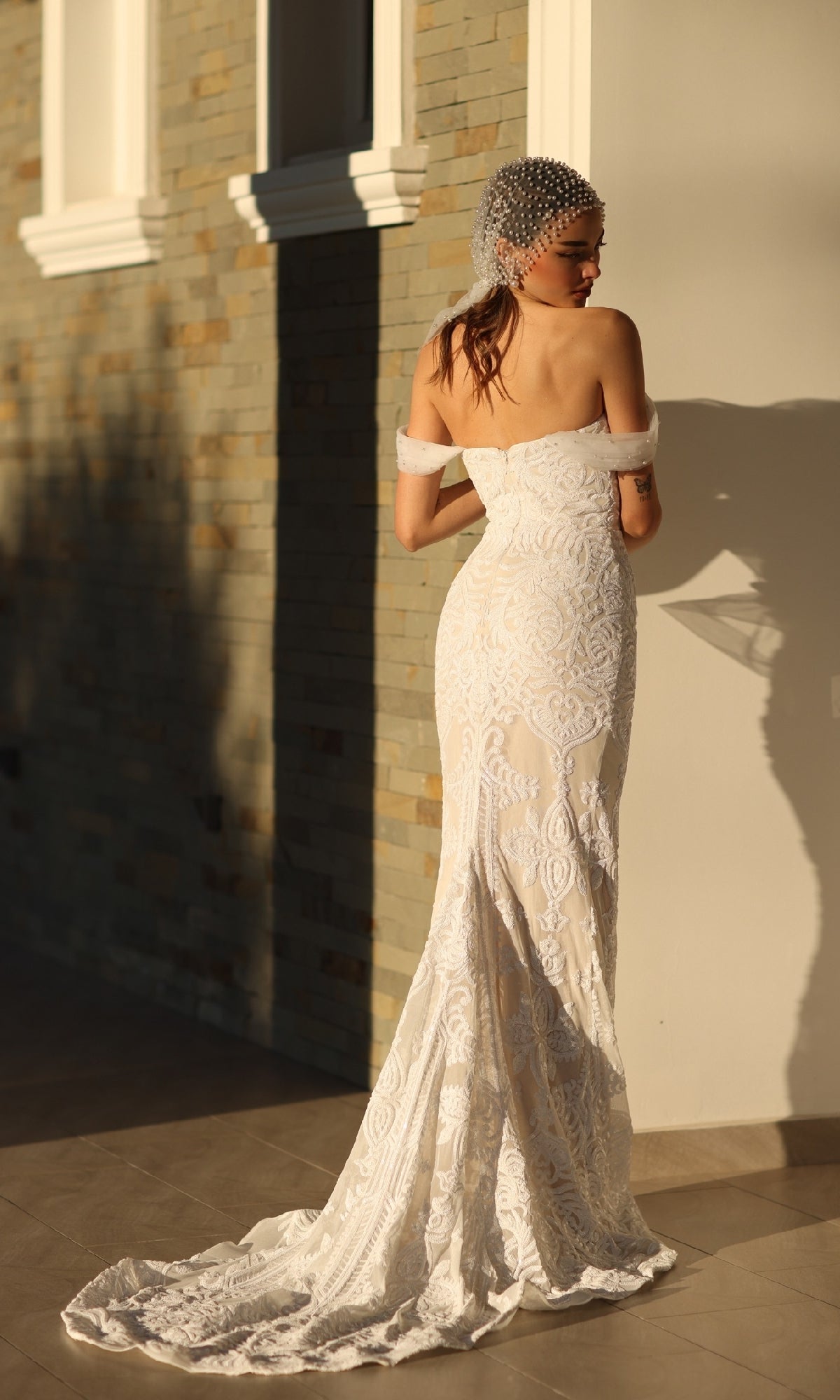 A woman in the Off-Shoulder White Bridal Gown: Tina Holly TW103 stands facing a wall as sunlight casts soft shadows and her beaded headpiece sparkles in the light.