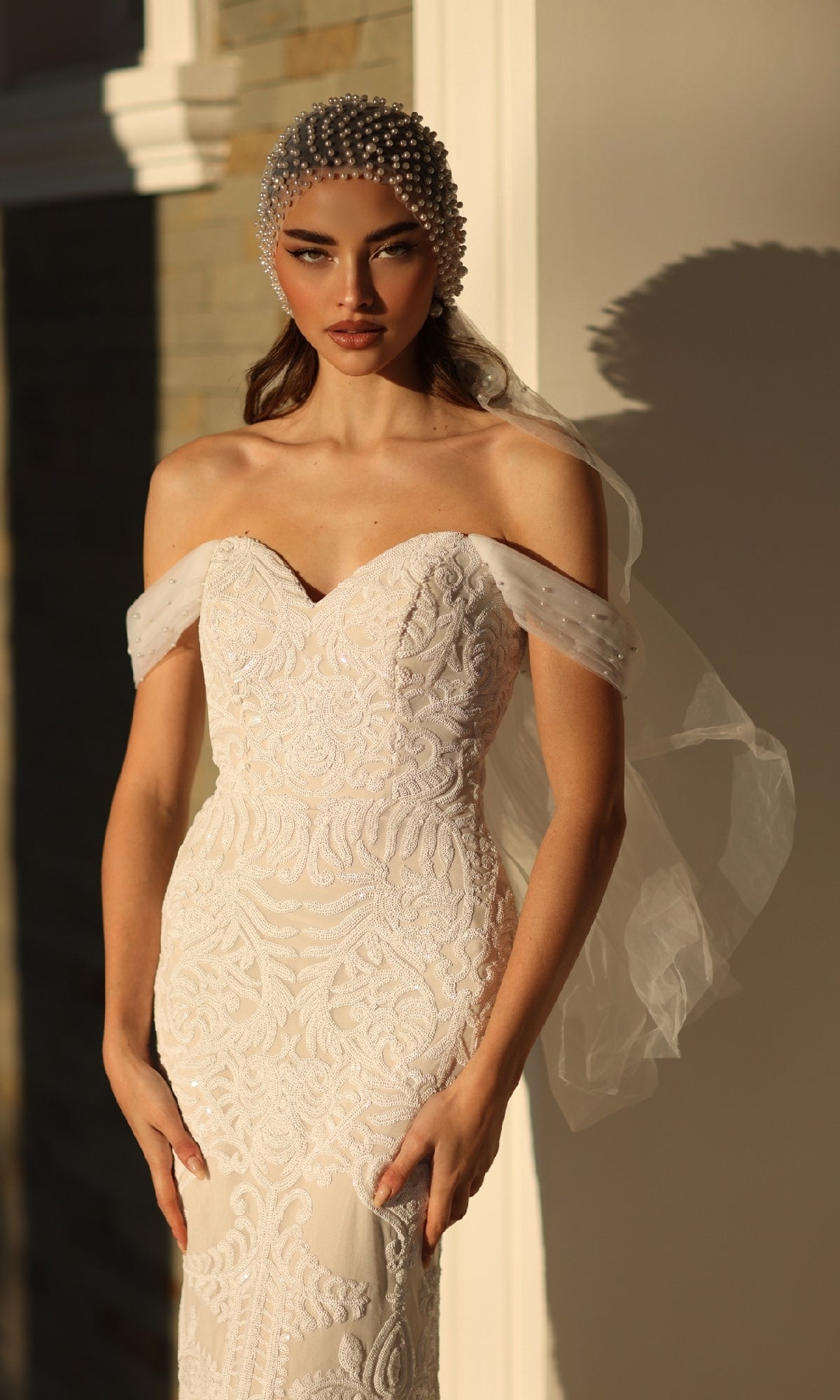 A woman in the Off-Shoulder White Bridal Gown: Tina Holly TW103 with fitted lace and a pearl headpiece poses gracefully against a sunlit wall.
