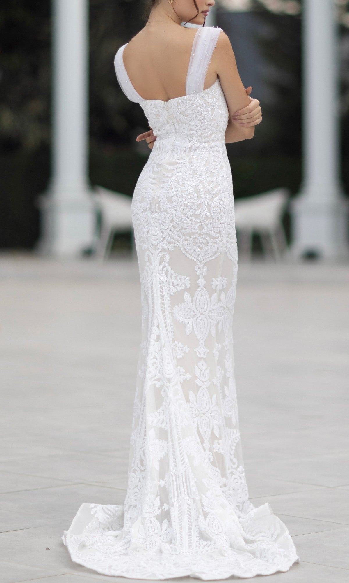 A woman stands outside seen from the back, arms crossed, wearing the fitted, floor-length Off-Shoulder White Bridal Gown: Tina Holly TW103 with intricate lace patterns.