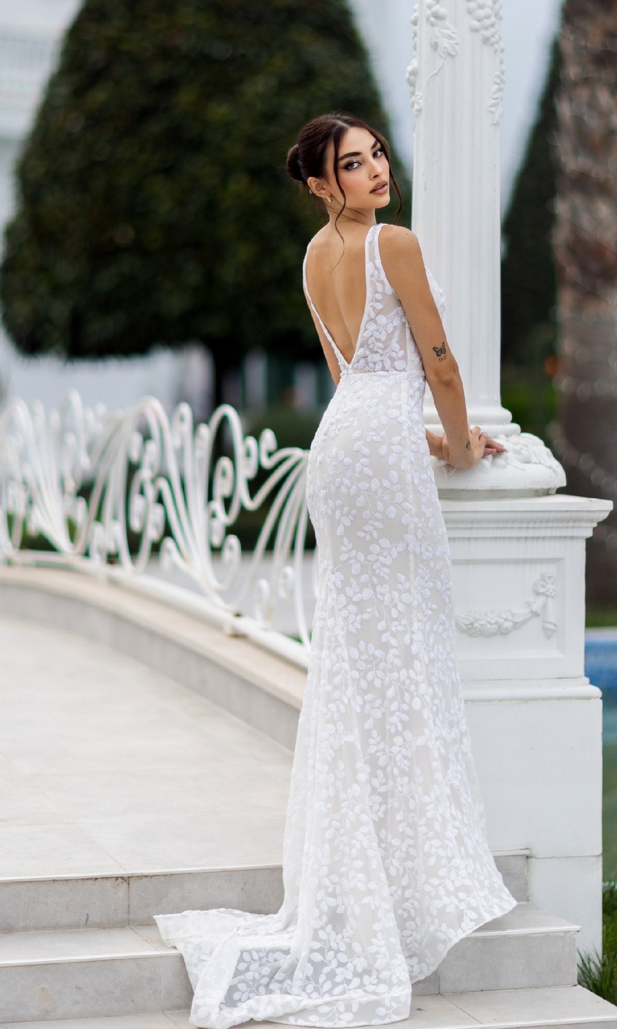 Wearing the Tina Holly TW108, a long white sequin formal dress with a sleeveless, backless design, a woman stands by a decorative column and looks over her shoulder.