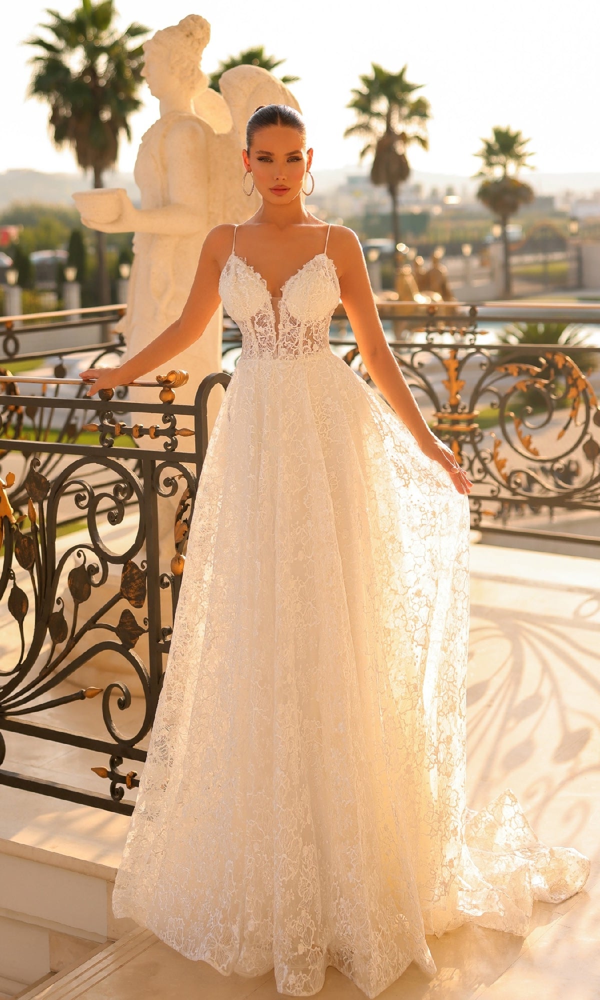 A woman in the Traditional Lace Bridal Gown: Tina Holly TW133, an off-white embroidered lace dress with thin straps, stands on a balcony adorned with ornate railings, palm trees, and statues in the background.