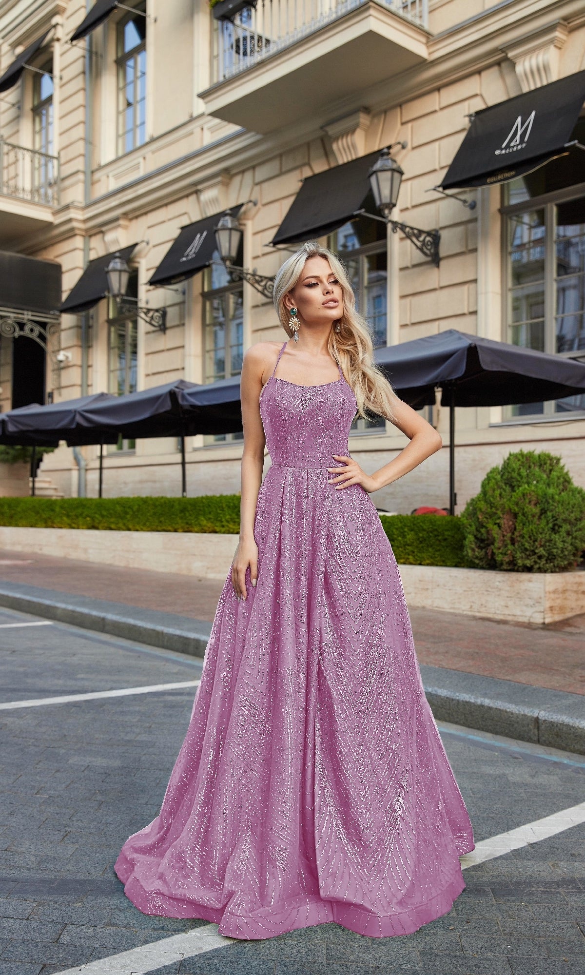 A woman poses outdoors on a city street in front of a beige building with black awnings, wearing the Backless Long Glitter Prom Dress: Tina Holly TW325.
