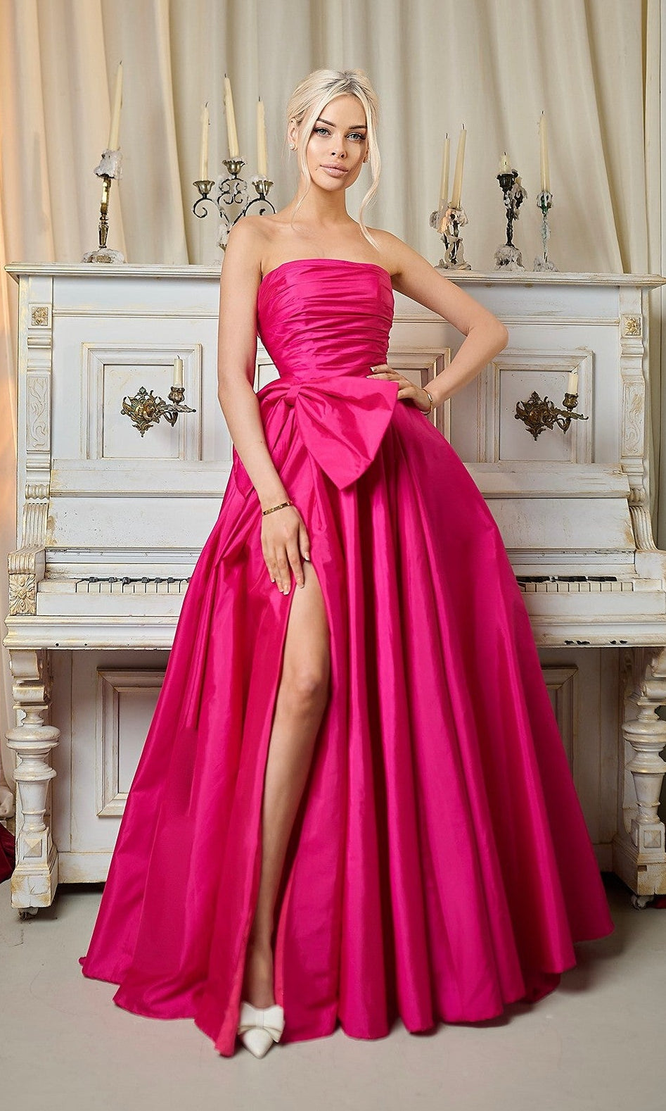 A woman stands before a white piano, wearing the Tina Holly TW603—an elegant strapless taffeta gown with ruched bodice, thigh-high slit, detachable bow, and pointed-toe heels. Candles and decor adorn the piano.