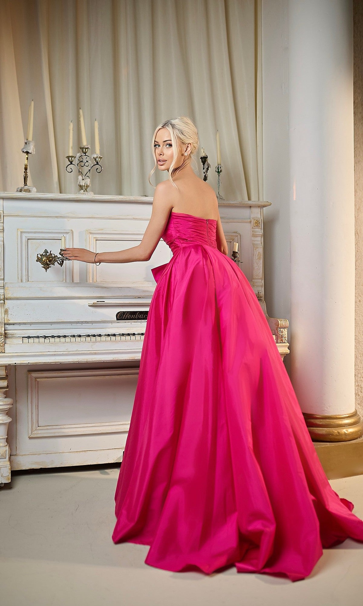A woman in the Long Formal Dress: Tina Holly TW603, featuring a strapless taffeta design with a ruched fitted bodice, stands by an ornate white piano, glancing over her shoulder in an elegant setting.