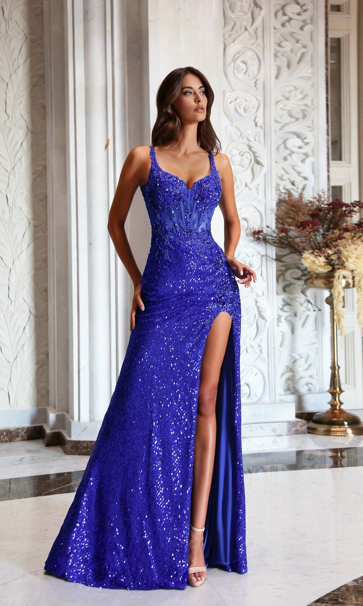 A woman wearing the Tina Holly TW612 long prom dress in royal blue, featuring a sleeveless design and high leg slit, stands indoors near ornate white walls and a decorative vase.