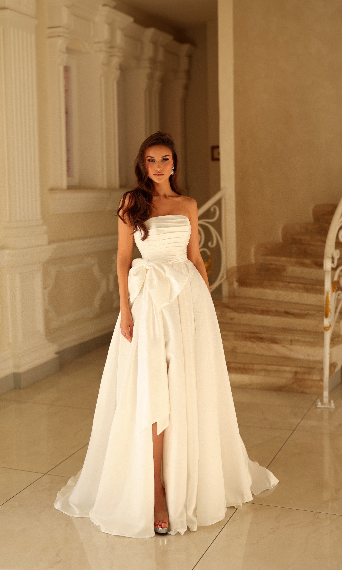 The Tina Holly TW621 long wedding dress in off-white features a strapless, floor-length silhouette with a ruched bodice and a large bow at the waist.