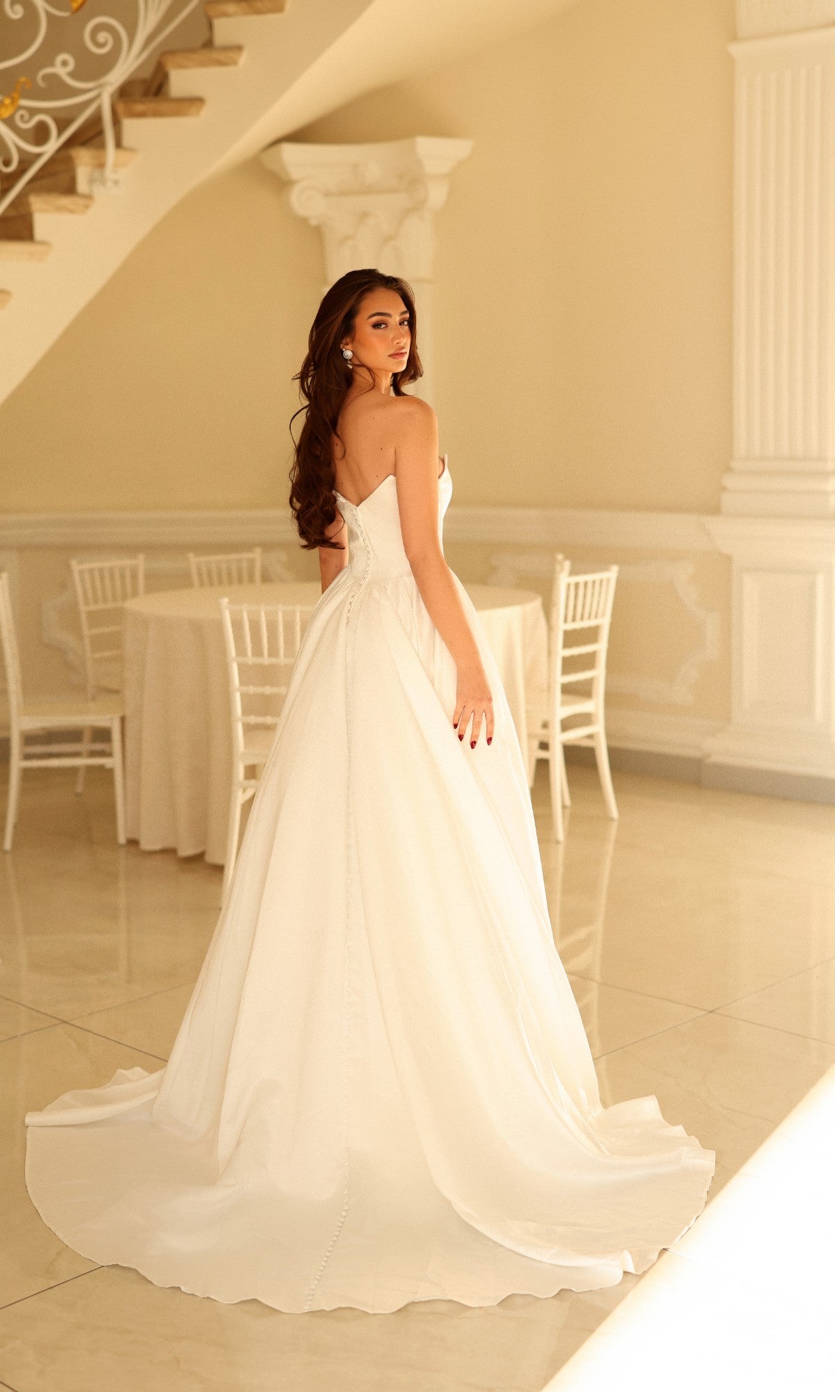 A woman wearing a Tina Holly TW622 long wedding dress, featuring a strapless A-line silhouette with a floor-length train, looking over her shoulder.