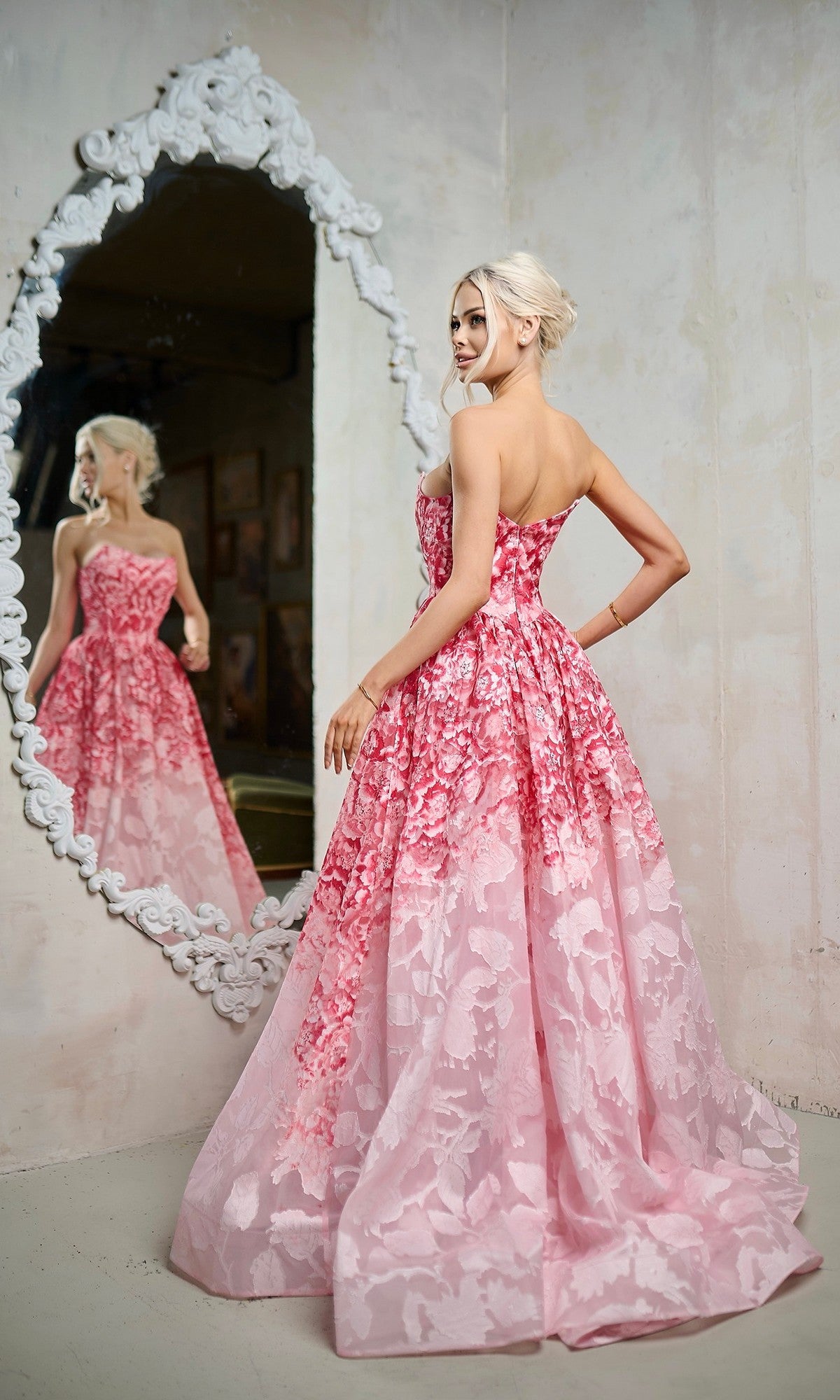 A blonde woman in the Tina Holly TW624 long formal dress stands before an ornate mirror, gazing at her reflection. The strapless A-line silhouette and soft pink floral organza jacquard fabric enhance her elegant pose.