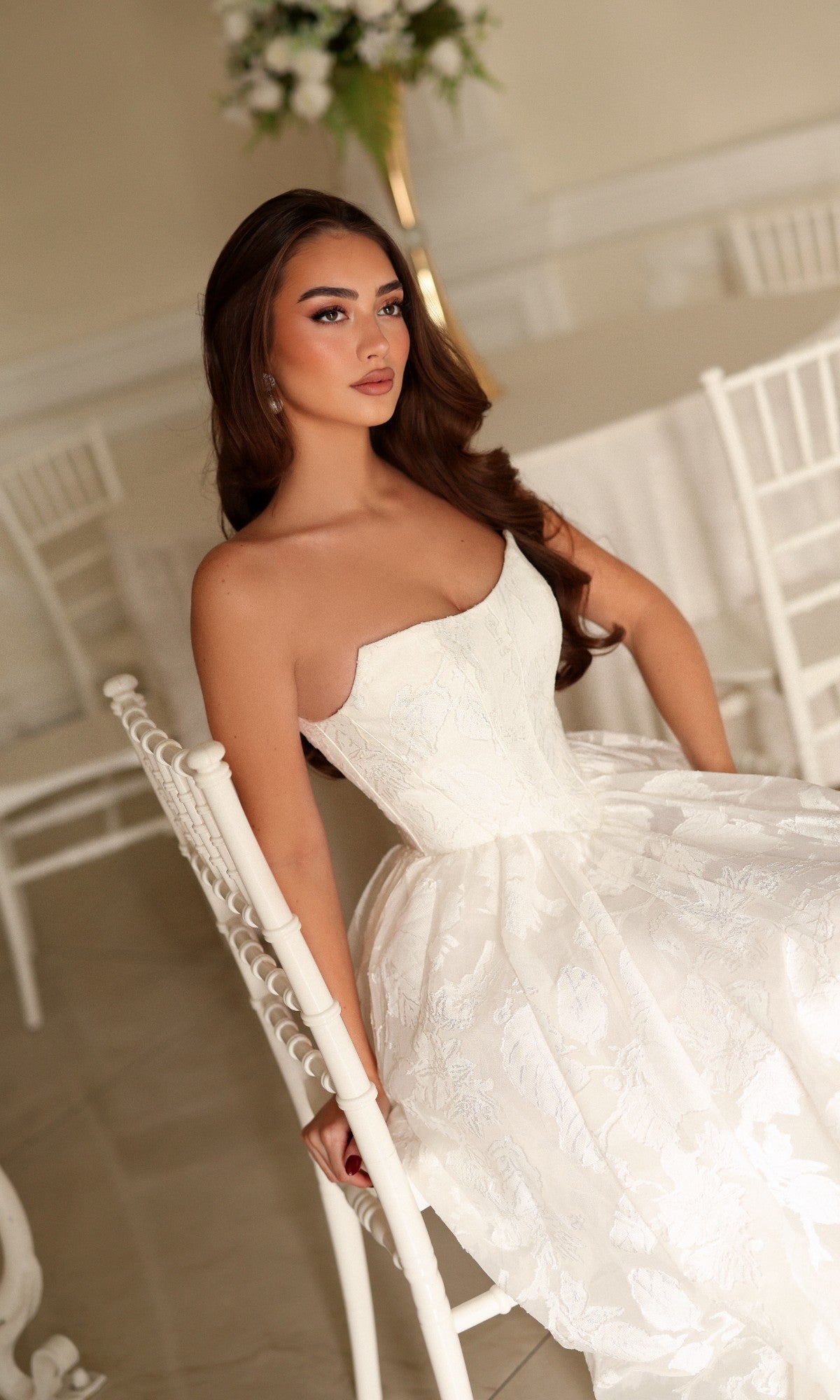 A woman wearing a Tina Holly TW627 long wedding dress with a strapless structured bodice and a full, floral-patterned organza jacquard skirt while seated on a white chair.