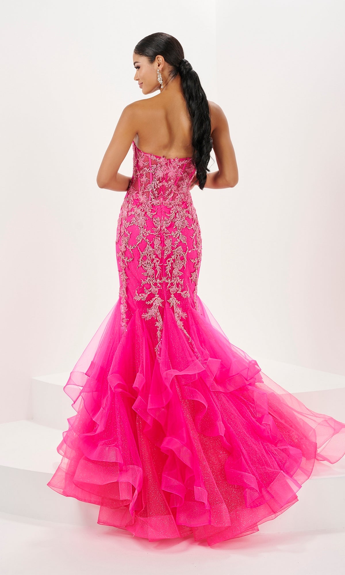 A woman stands on white steps, facing away from the camera, wearing the Long Prom Dress 16057 by Tiffany—a strapless mermaid gown with a sweetheart neckline and gold embroidery.