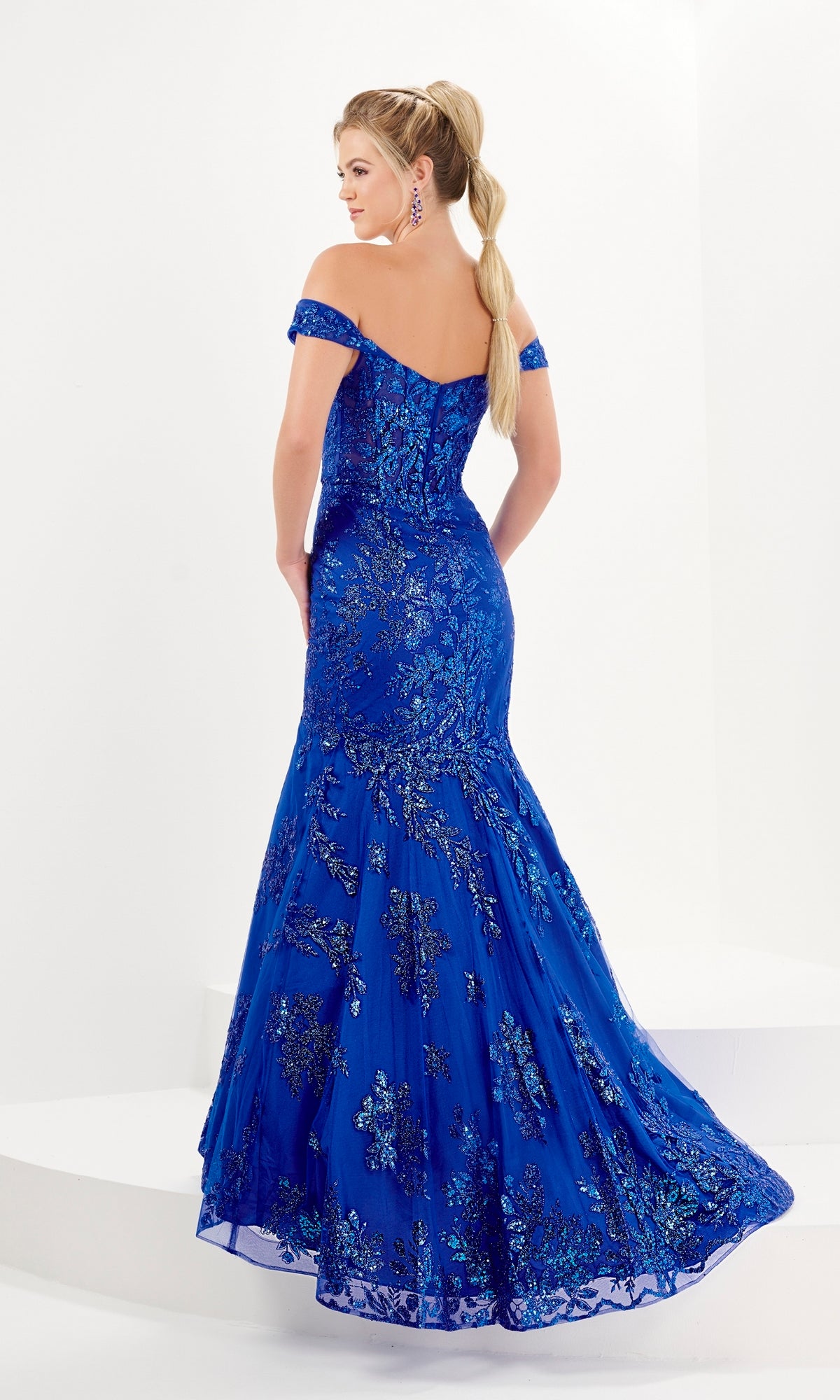 Woman in the Tiffany Sheer Corset Glitter Mermaid Prom Dress 16080, featuring a bright blue off-the-shoulder style with sparkling embellishments and a sheer corset bodice, stands against a plain white background, facing away from the camera.