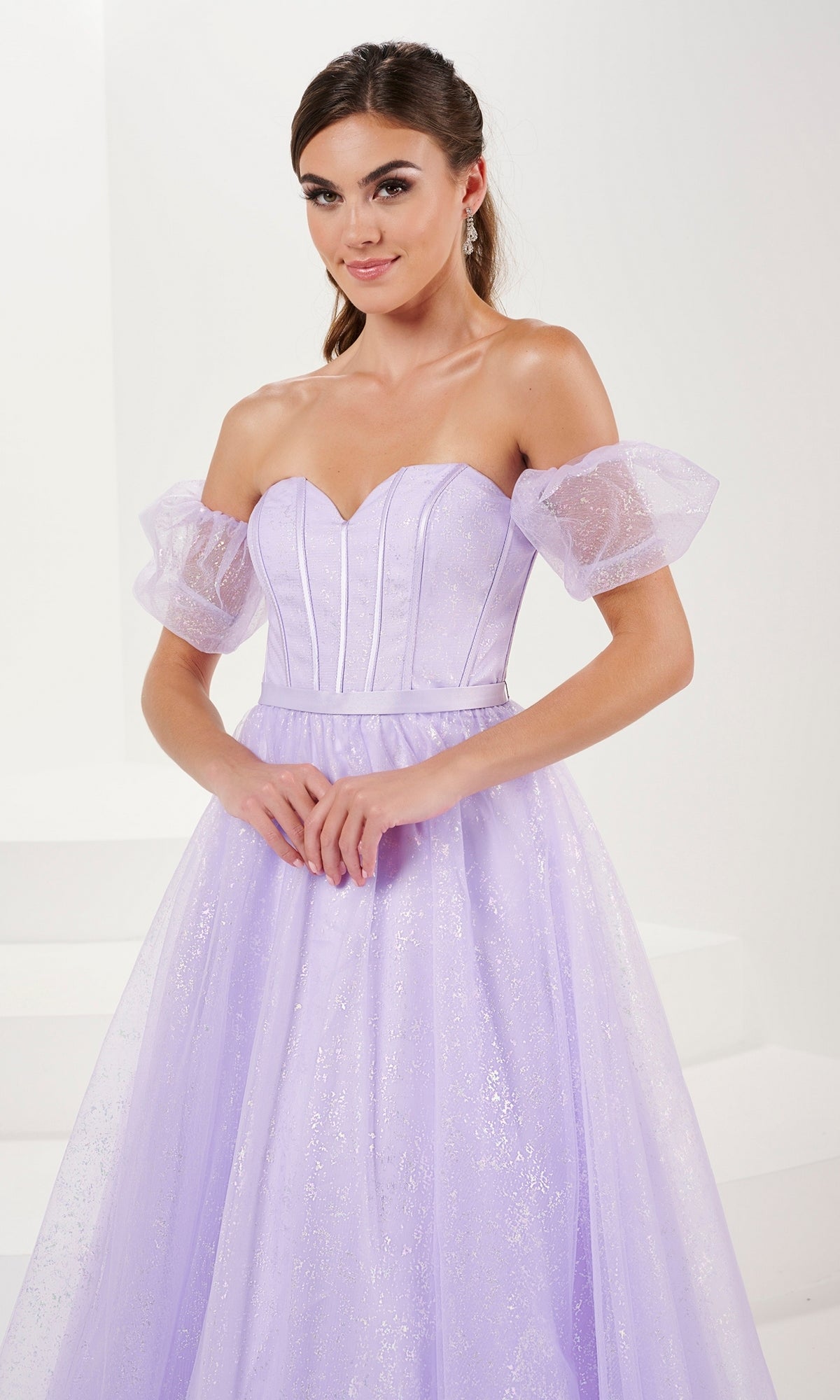 A woman wears the Tiffany Strapless Glitter Pastel Prom Dress 16083—a light purple, glitter tulle ball gown with a corset bodice—standing against a plain white background.